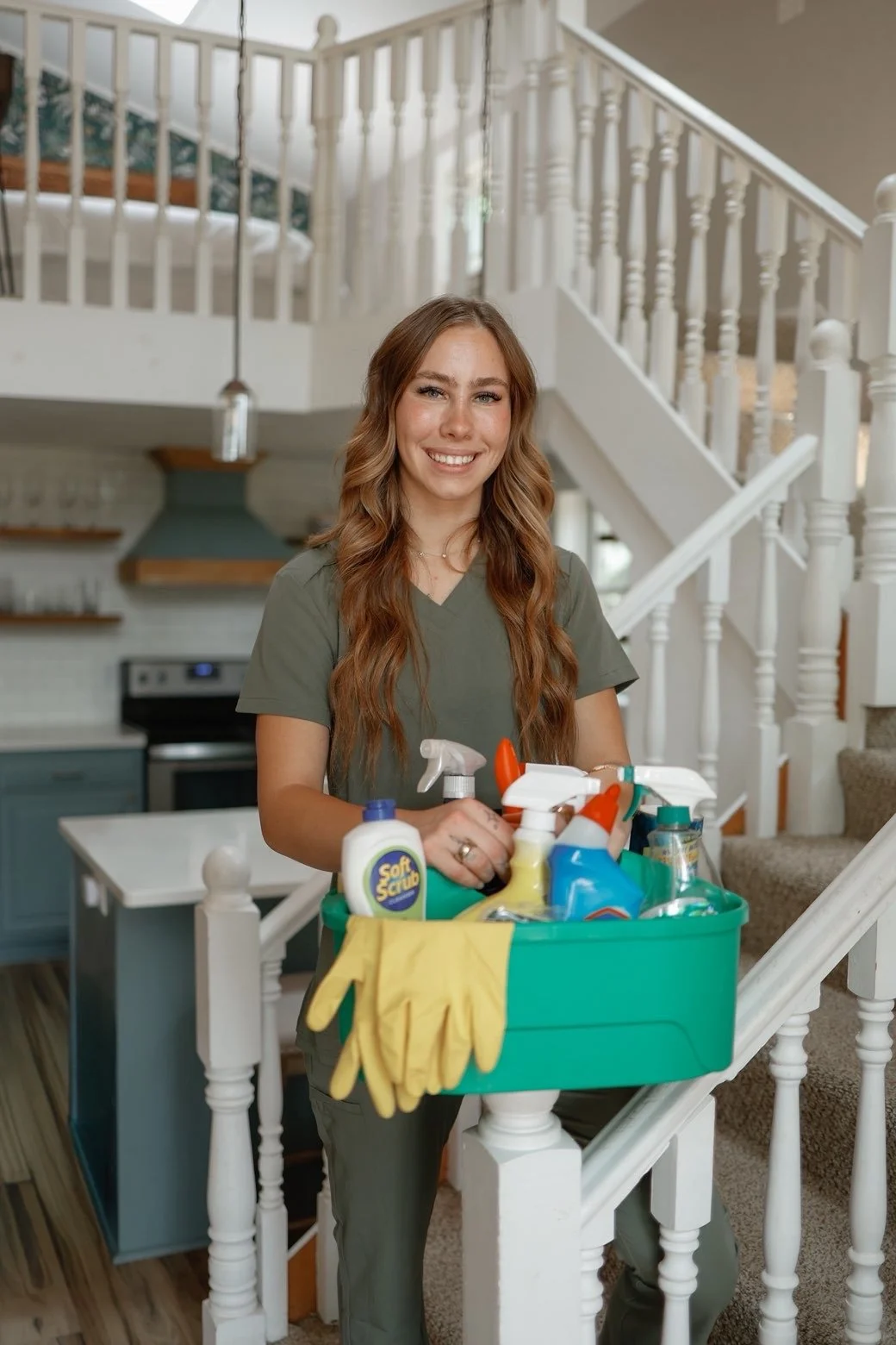 A woman with long, wavy brown hair, smiling, holding a cleaning caddy with various cleaning supplies including spray bottles, gloves, and a bottle of soft scrub, standing on a staircase in a home.