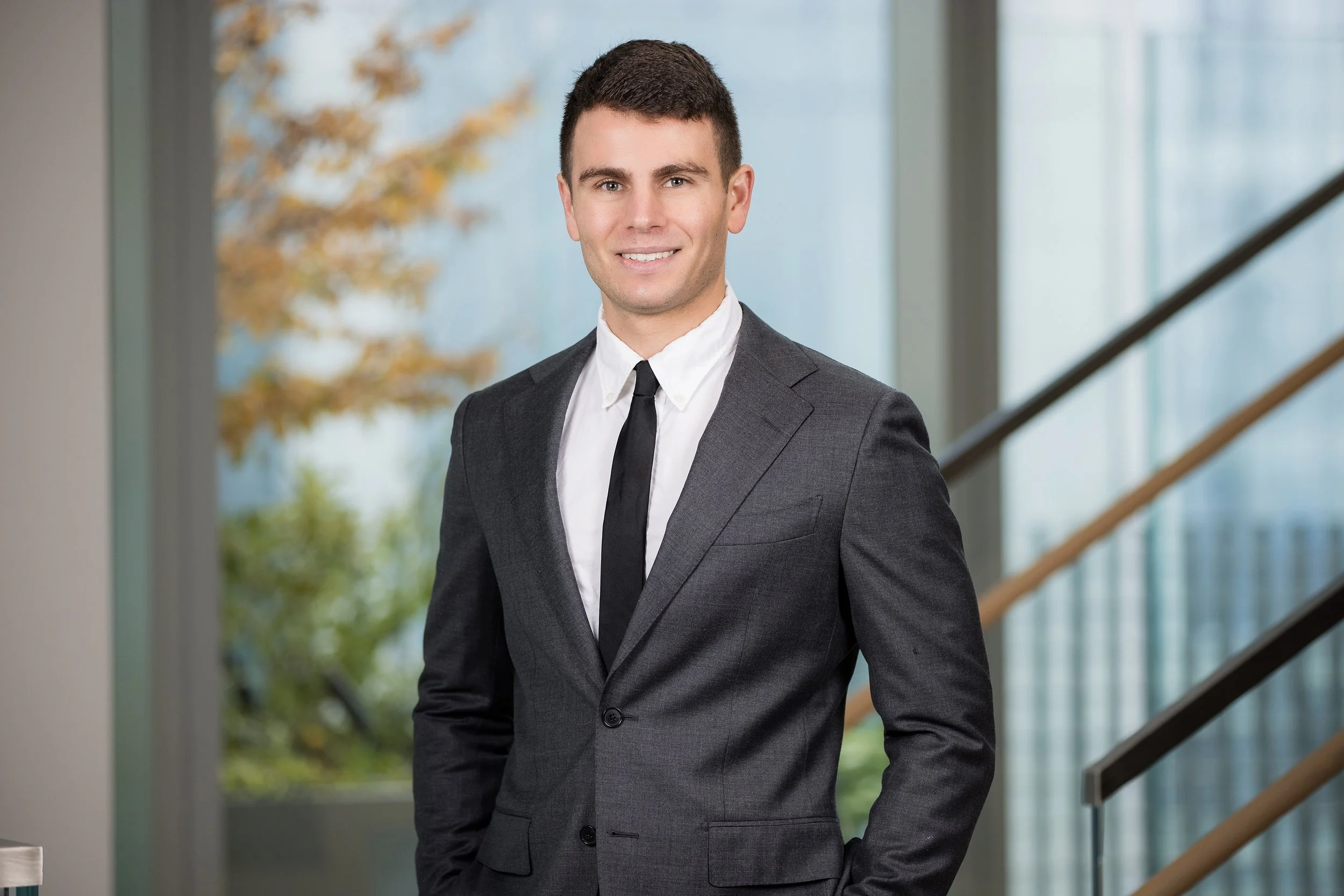 A young man in a gray suit with a white shirt and black tie, smiling and standing indoors near a staircase with large windows and blurred outdoor scenery in the background.