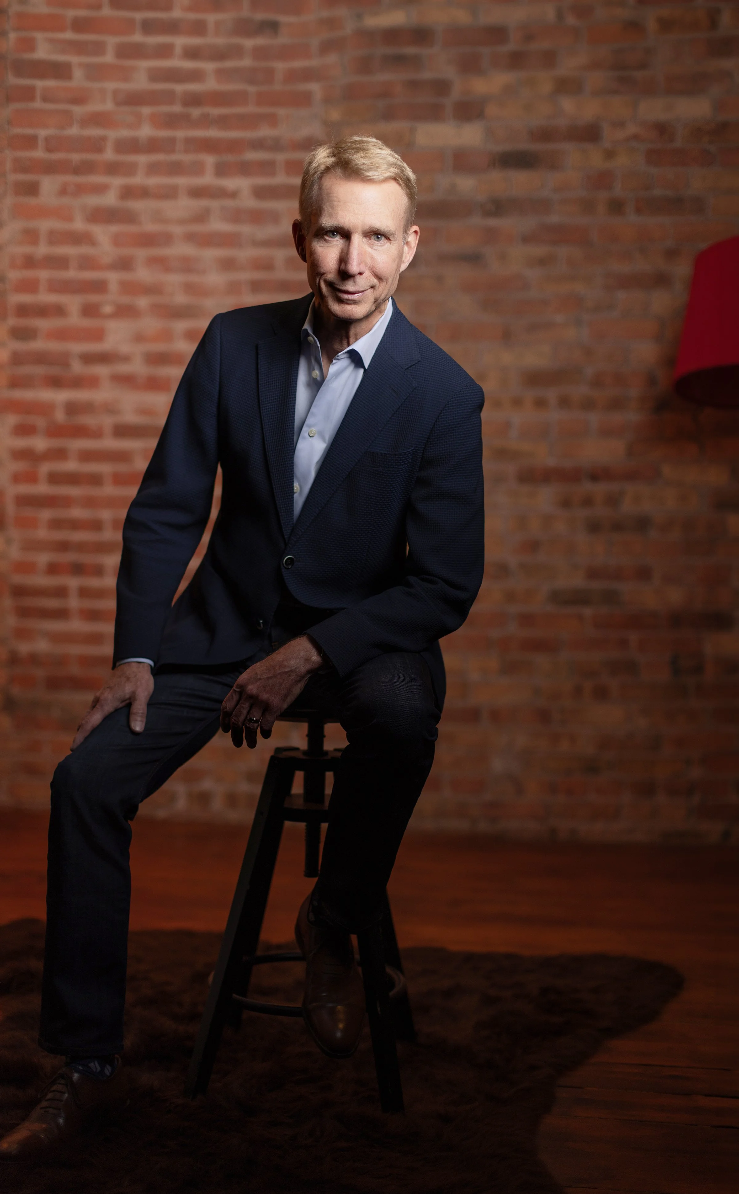 A middle-aged man with blond hair in a blue suit jacket and light blue shirt sitting on a high stool in front of a brick wall background.
