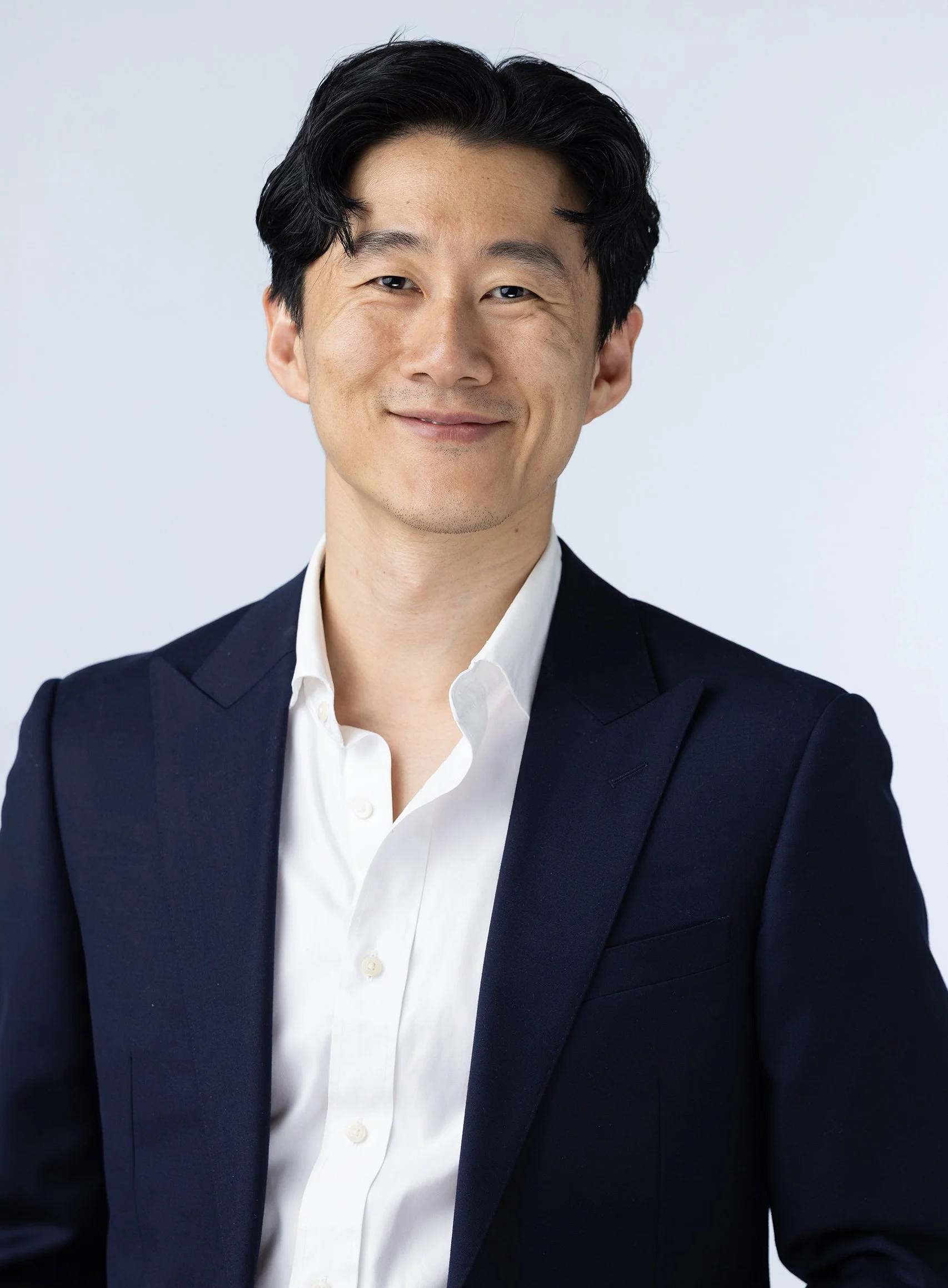 Portrait of a smiling man with dark hair wearing a white shirt and a dark suit jacket against a plain light background.