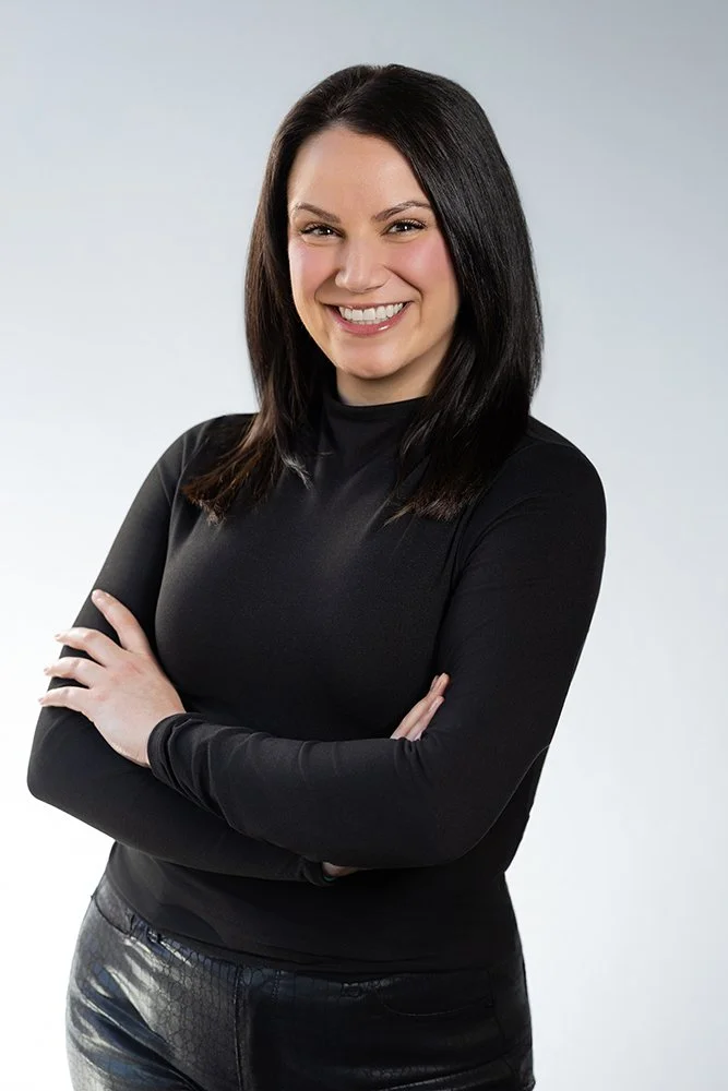 A woman with long dark hair smiling, wearing a black long-sleeve shirt and black leather pants, standing with arms crossed against a gradient gray background.
