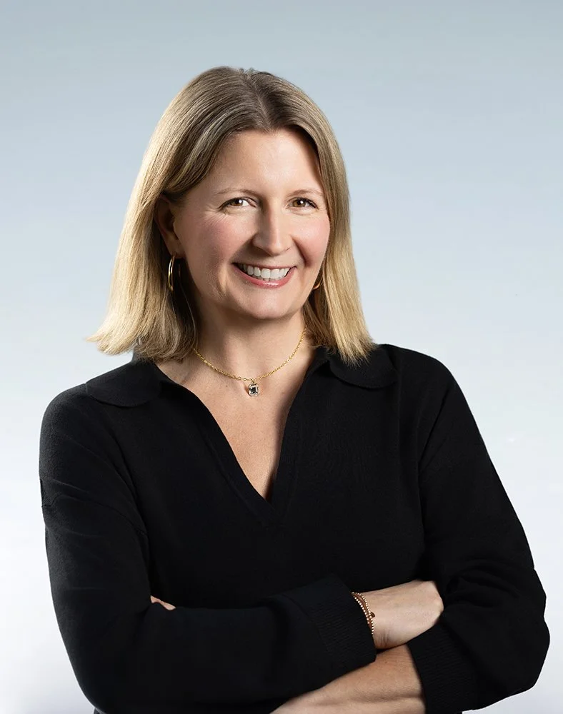 A woman with shoulder-length blonde hair, smiling, wearing a black collared top, gold jewelry, and standing against a light blue background.