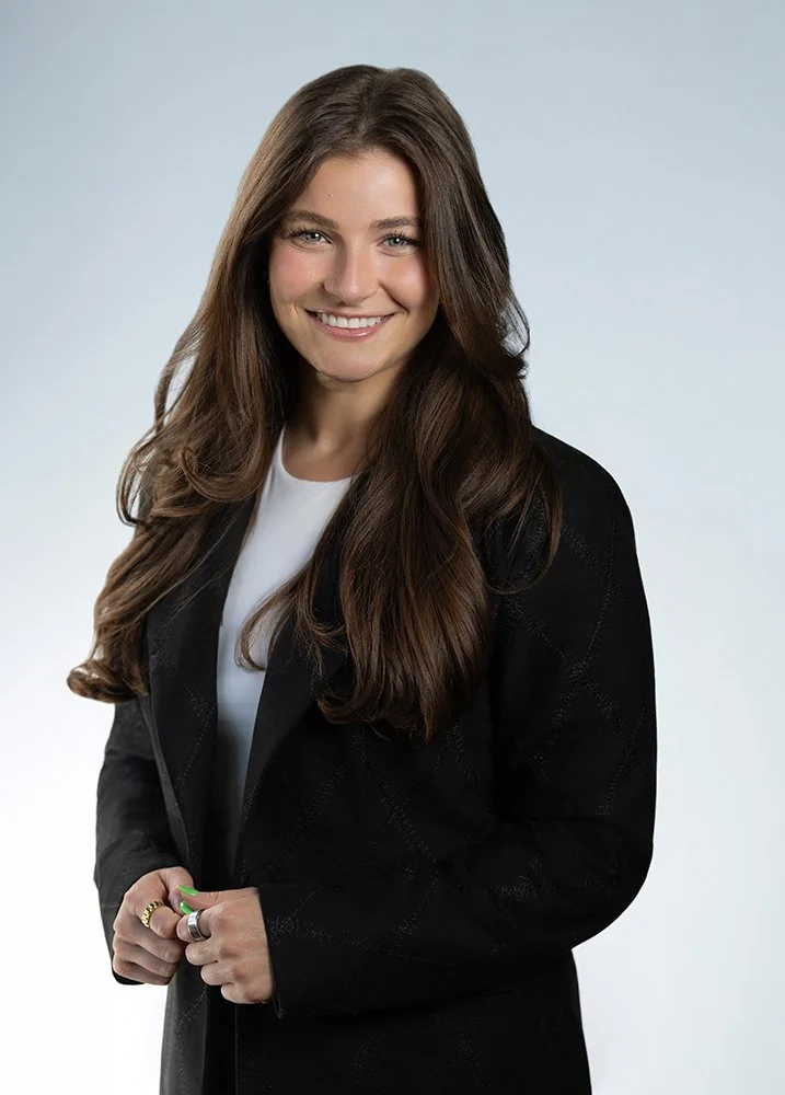 A woman with long brown hair and a big smile, wearing a black blazer over a white top, standing against a plain light gray background.