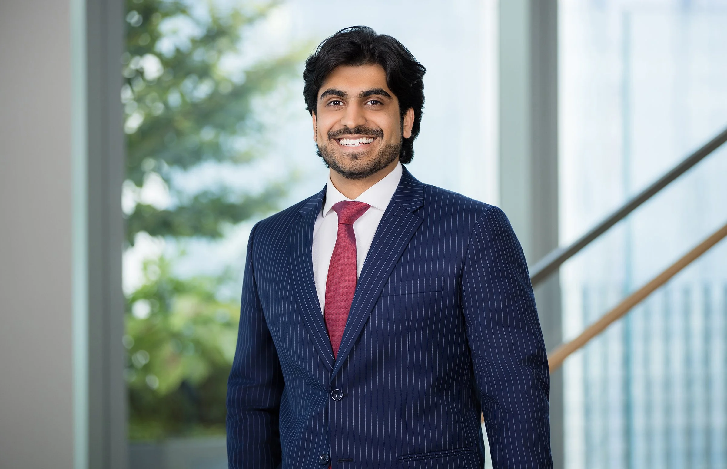 A young man with dark hair, a beard, and a bright smile, wearing a dark blue pinstripe suit and a red tie, standing in a modern building with large windows and green trees outside.