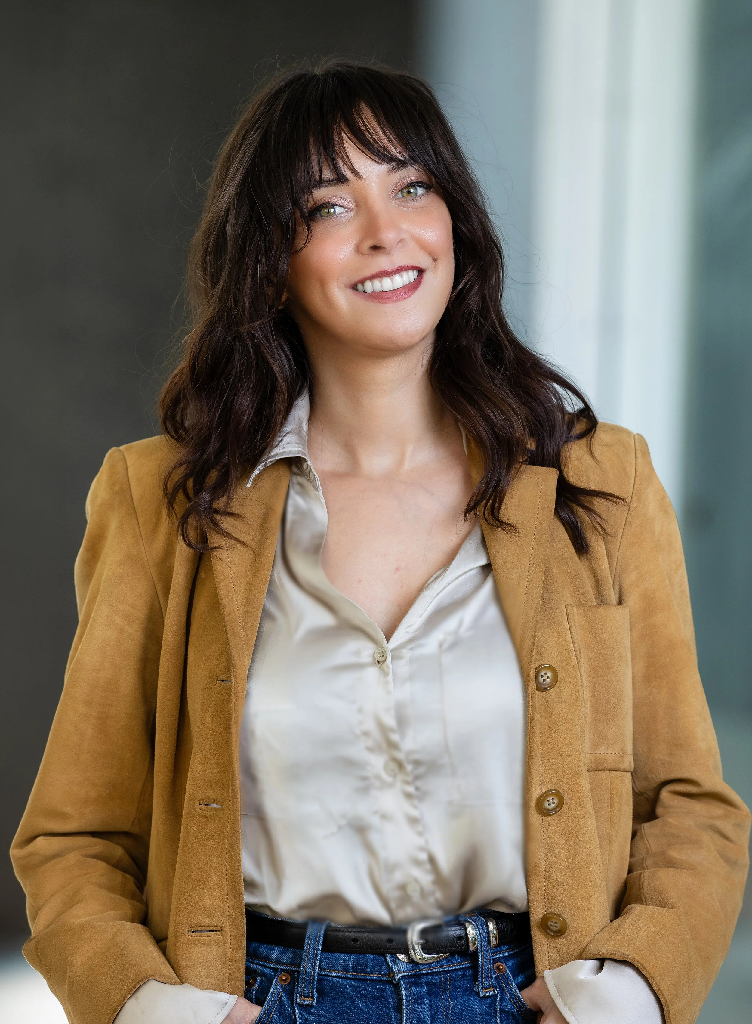 A woman with dark wavy hair, green eyes, and a bright smile wearing a light beige satin blouse, a tan blazer, and blue jeans.