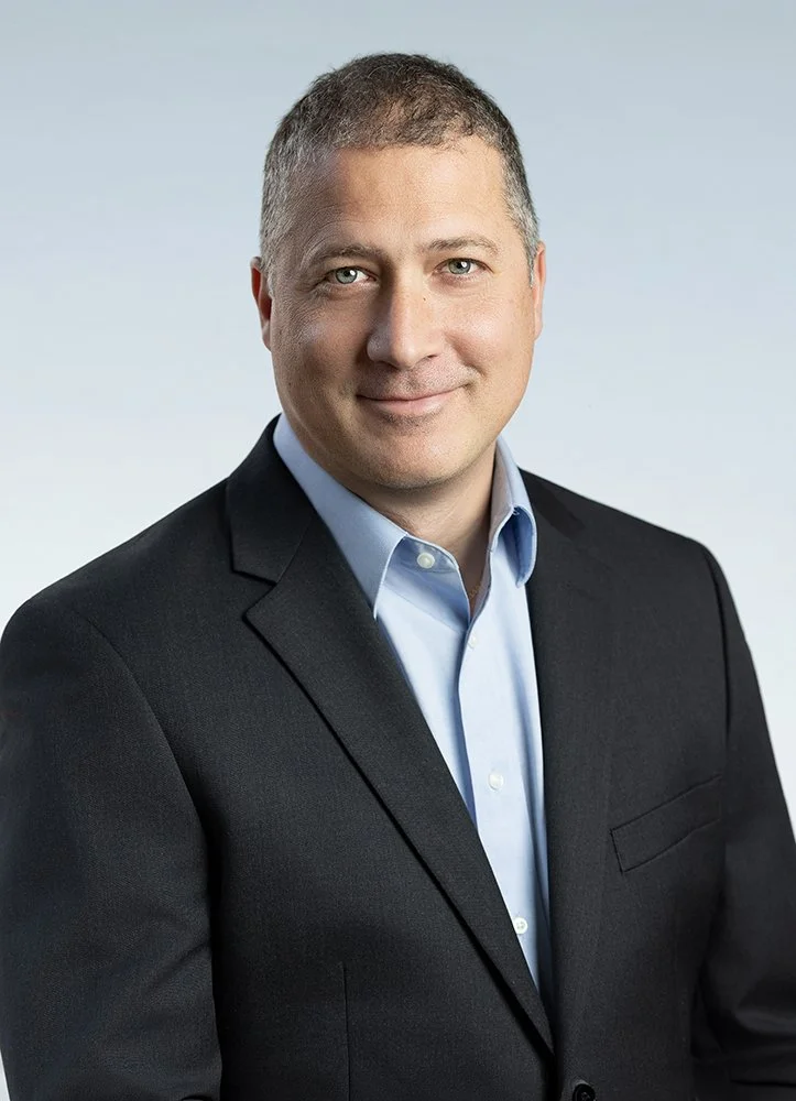 A man in a dark suit with a light blue dress shirt, smiling, against a plain light background.