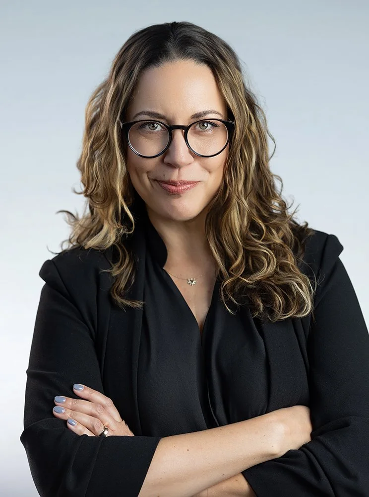 A woman with curly brown hair wearing glasses and a black blazer, smiling with arms crossed, against a plain light blue background.