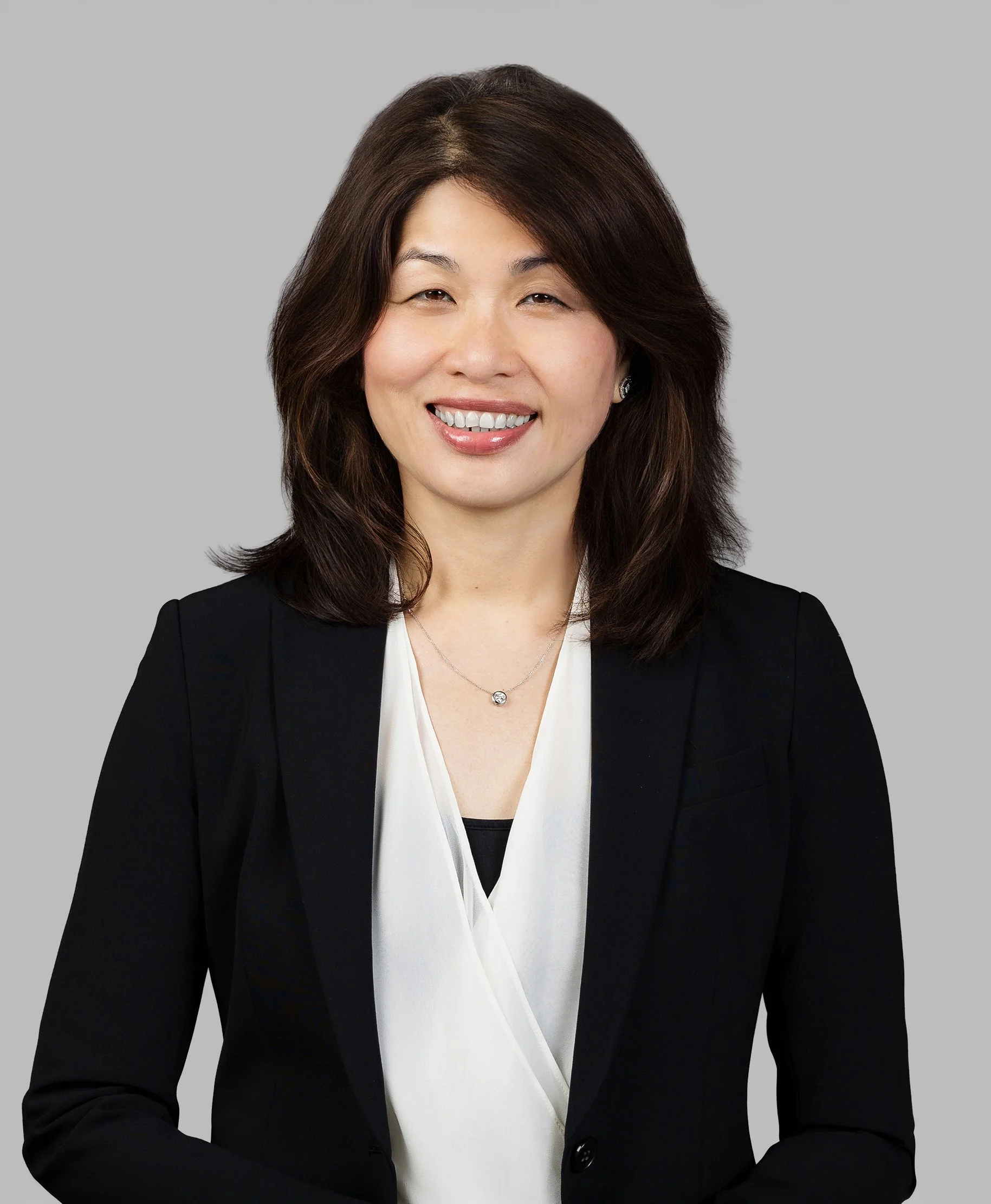 Headshot of an Asian woman with shoulder-length brown hair, wearing a black blazer over a white blouse, smiling against a light gray background.
