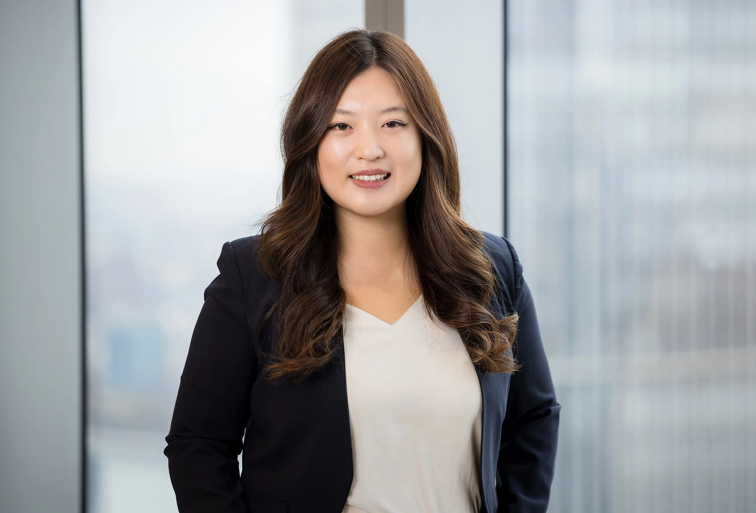 A woman with long, wavy brown hair wearing a navy blazer and white top, smiling, standing in front of a blurred modern office background.