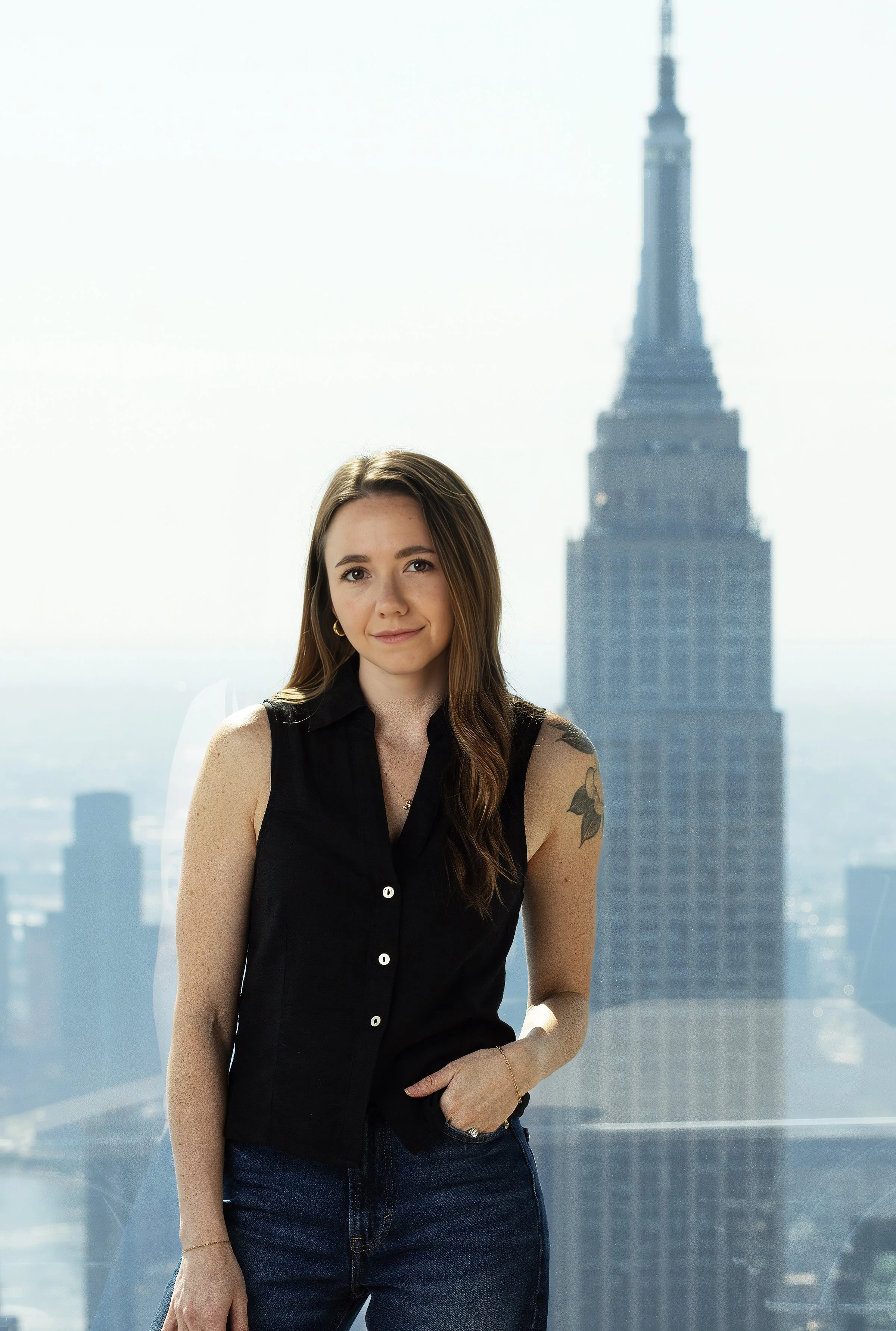 A woman with long brown hair and tattoos on her arm standing in front of a window with a cityscape and the Empire State Building in the background.
