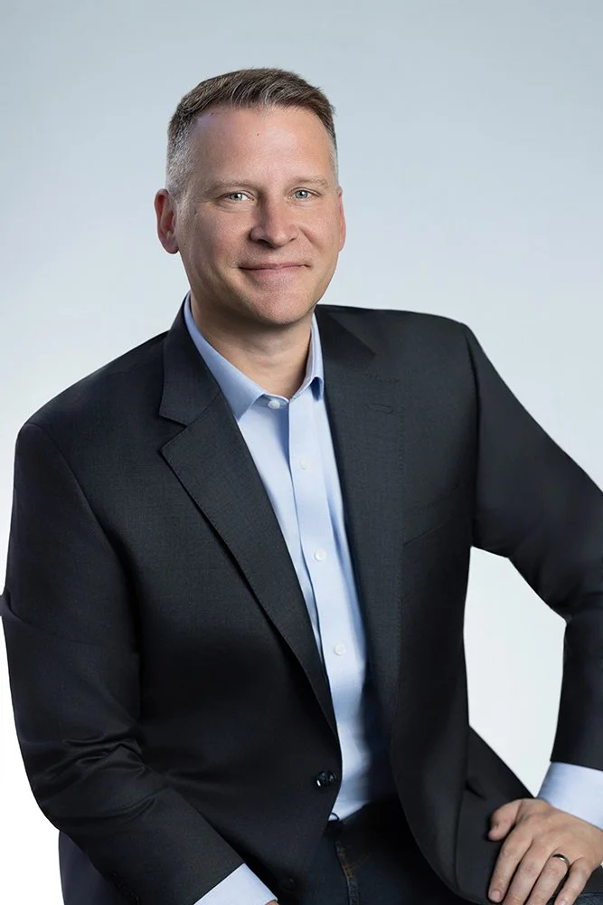 Professional portrait of a man in a dark suit with light blue shirt, smiling with a confident pose against a neutral background.
