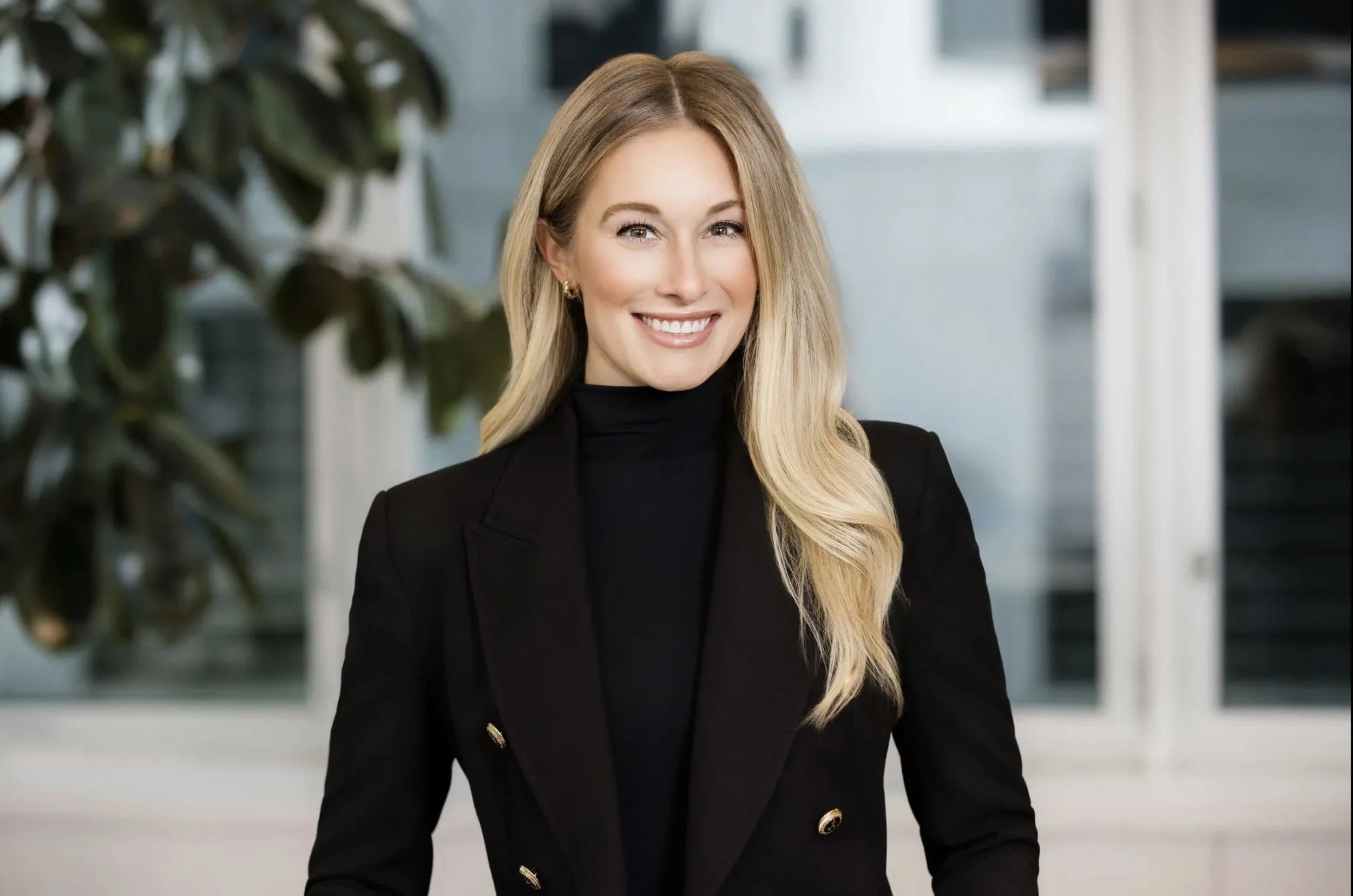 A woman with long blonde hair smiling in a professional setting, wearing a black blazer with gold buttons and a black turtleneck.