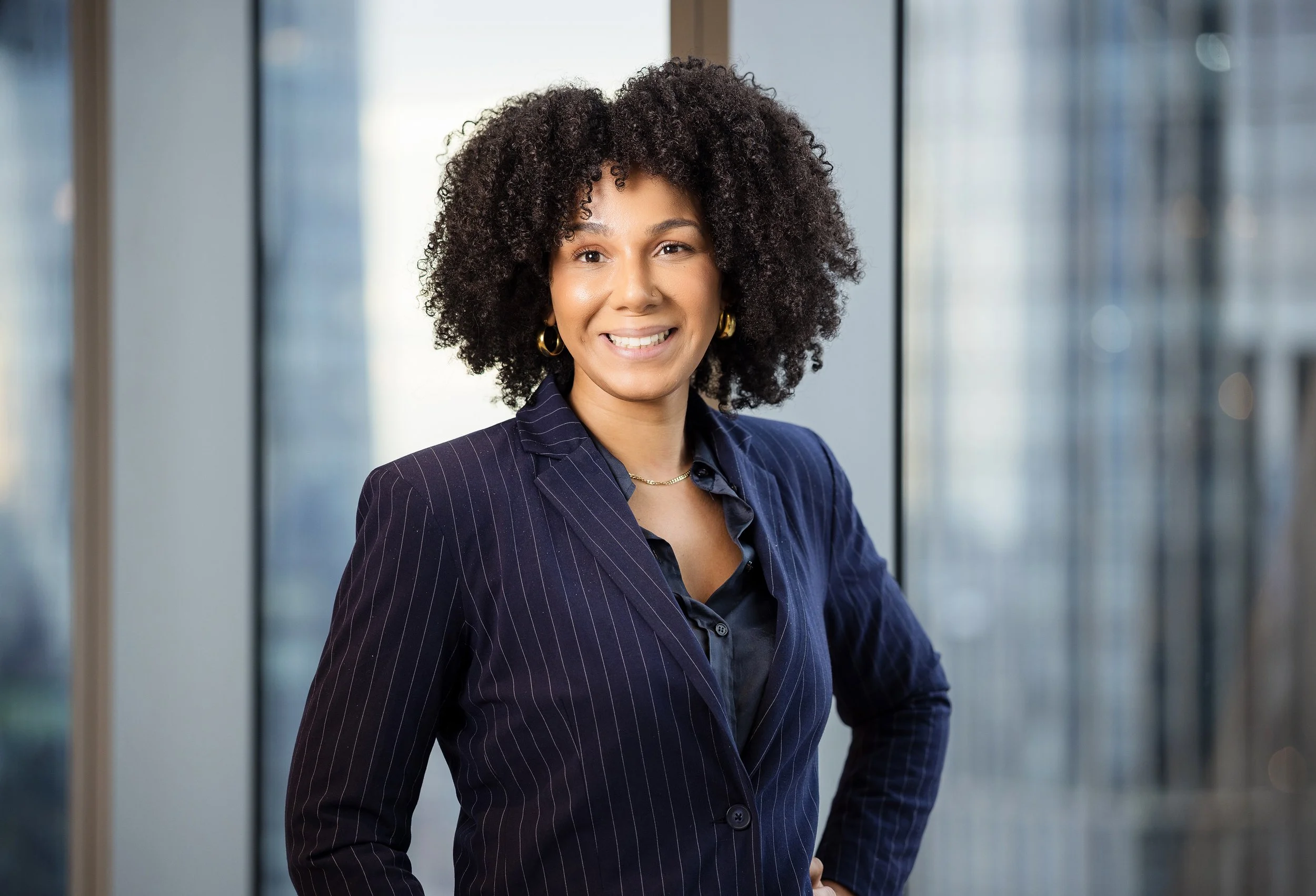 A woman with naturally curly hair wearing a pinstripe blazer and gold earrings, smiling at the camera indoors.