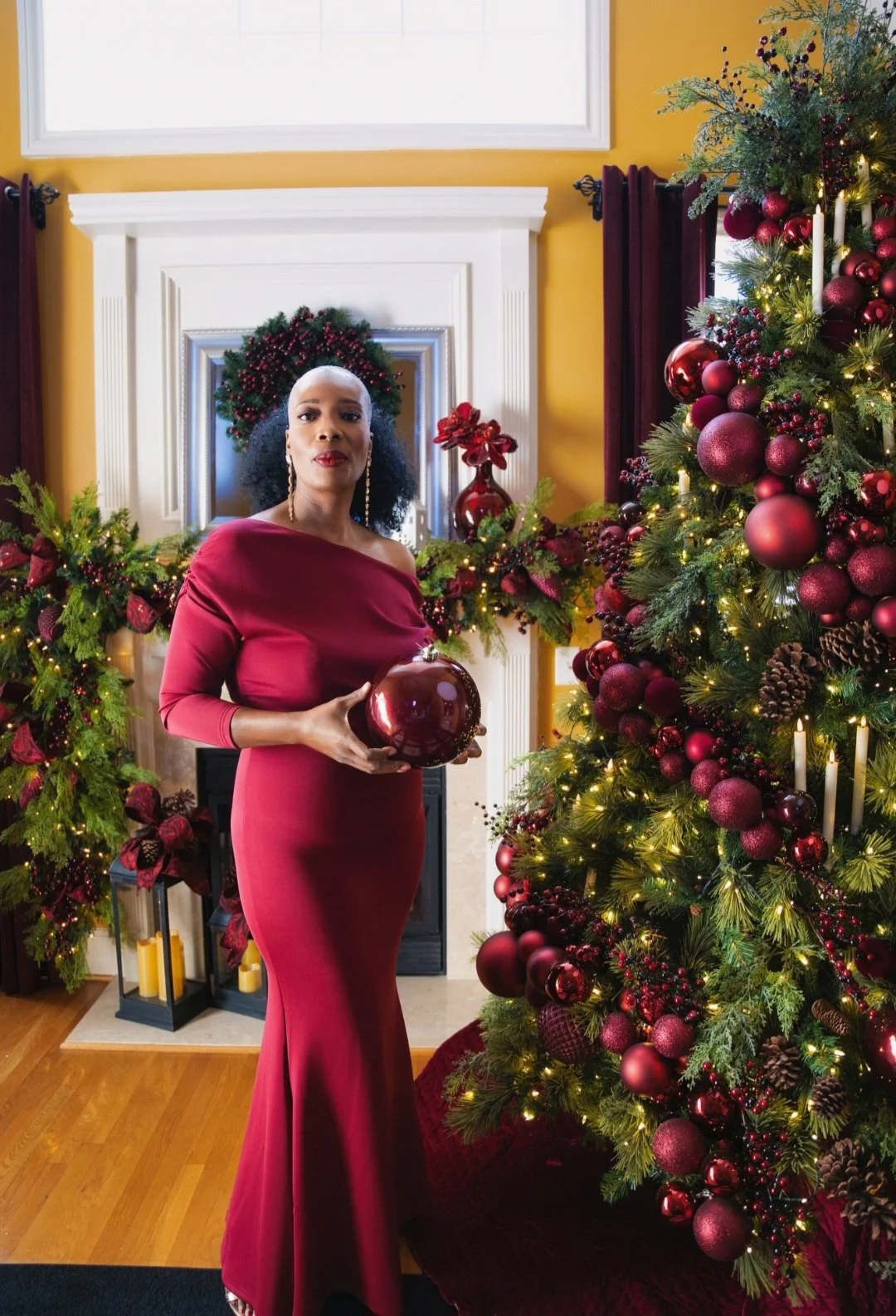 A woman in a red gown holding a large red Christmas ornament, standing in a decorated living room with a Christmas tree, wreath, and festive decorations.