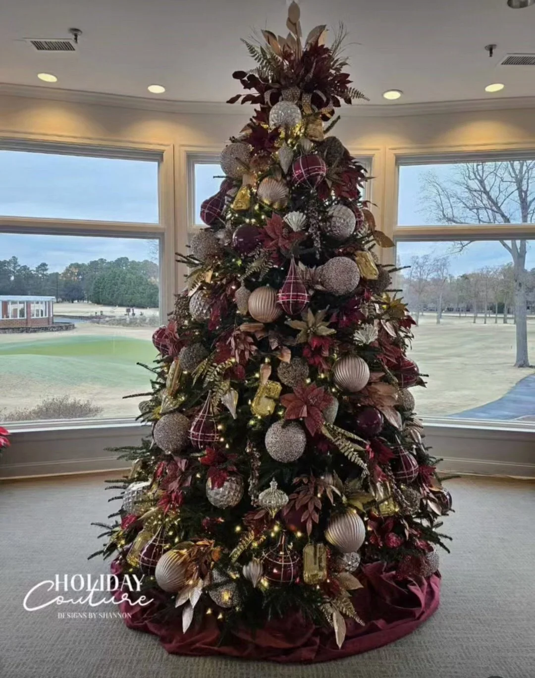 Decorated Christmas tree with gold, red, and silver ornaments inside a room with large windows showing an outdoor landscape.