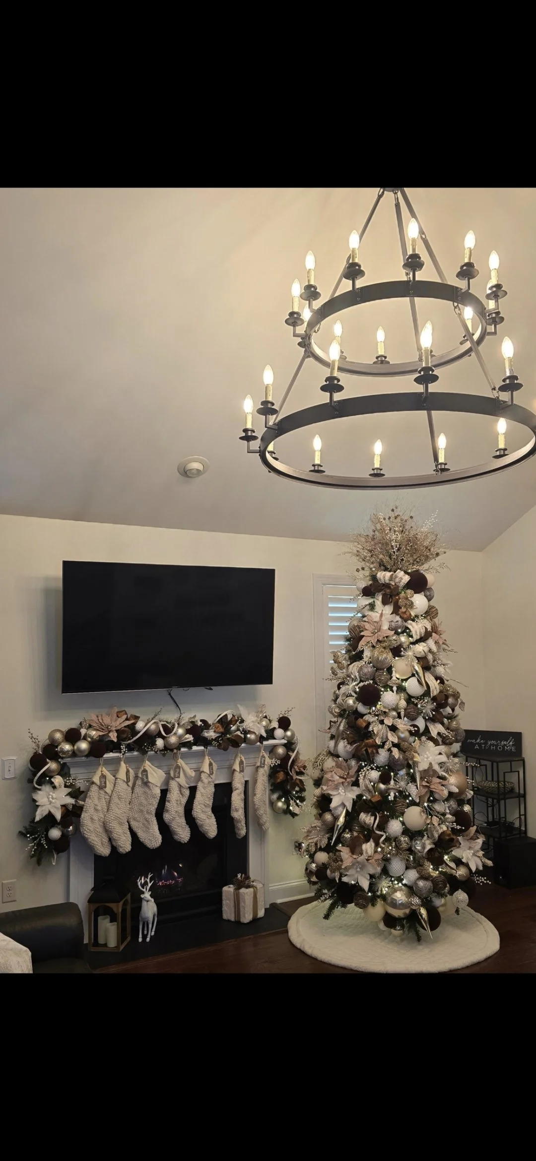 Living room decorated for Christmas with a tall decorated tree, stockings hanging on the fireplace, and a chandelier overhead.