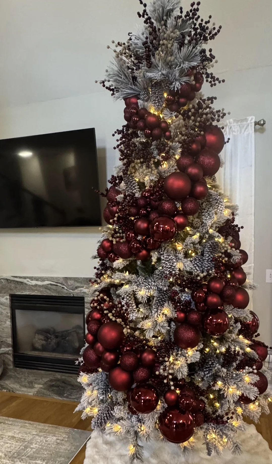 A decorated Christmas tree with white frosted branches, red ornaments, berries, pinecones, and yellow lights, positioned near a fireplace and a wall-mounted TV.