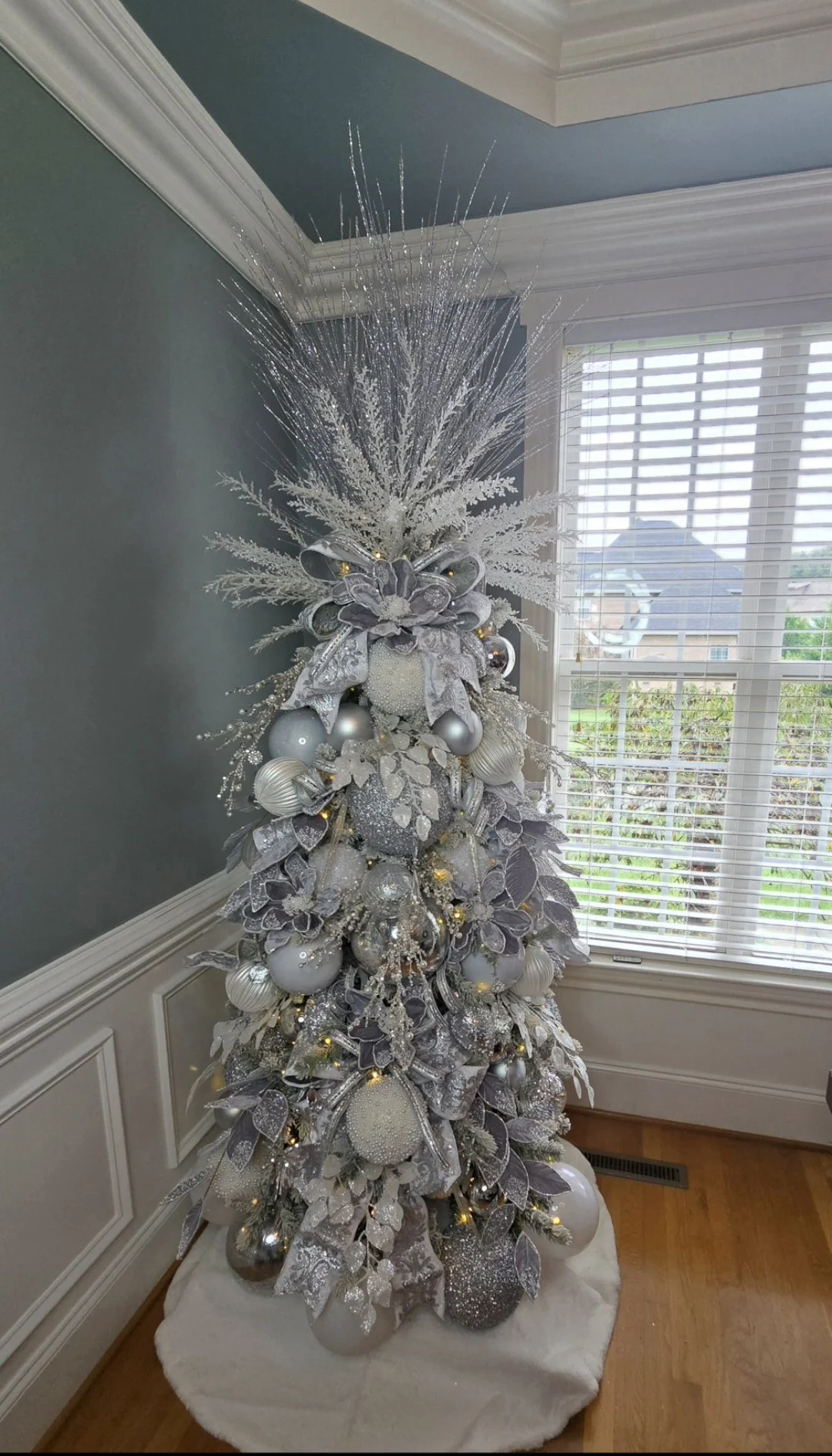 A Christmas tree decorated in silver and white ornaments, ribbons, and glitter, topped with a frosted branch arrangement, situated in a corner near a window with white blinds.
