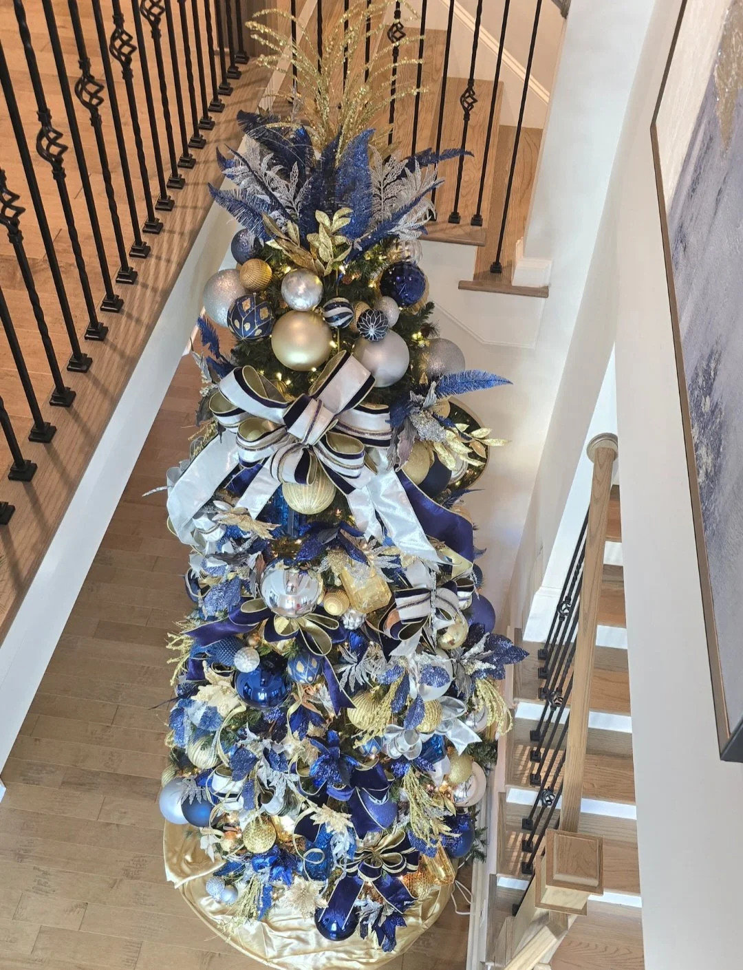 Decorated Christmas tree with blue, gold, and silver ornaments, ribbons, and large leaves, placed on a blue and gold tree skirt in a home foyer.