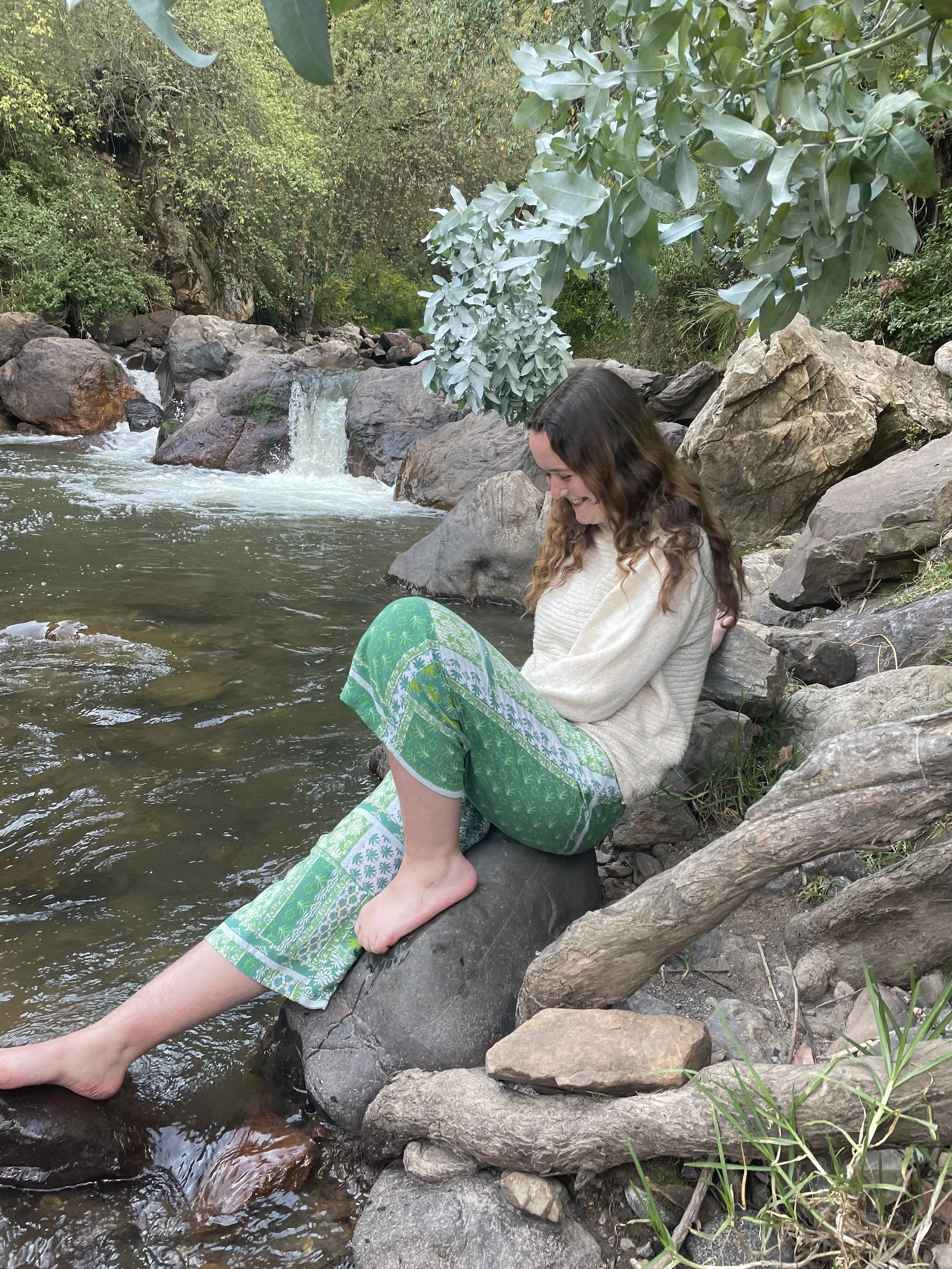 A woman sitting by a stream surrounded by rocks and green foliage, smiling with her feet dipped in the water.