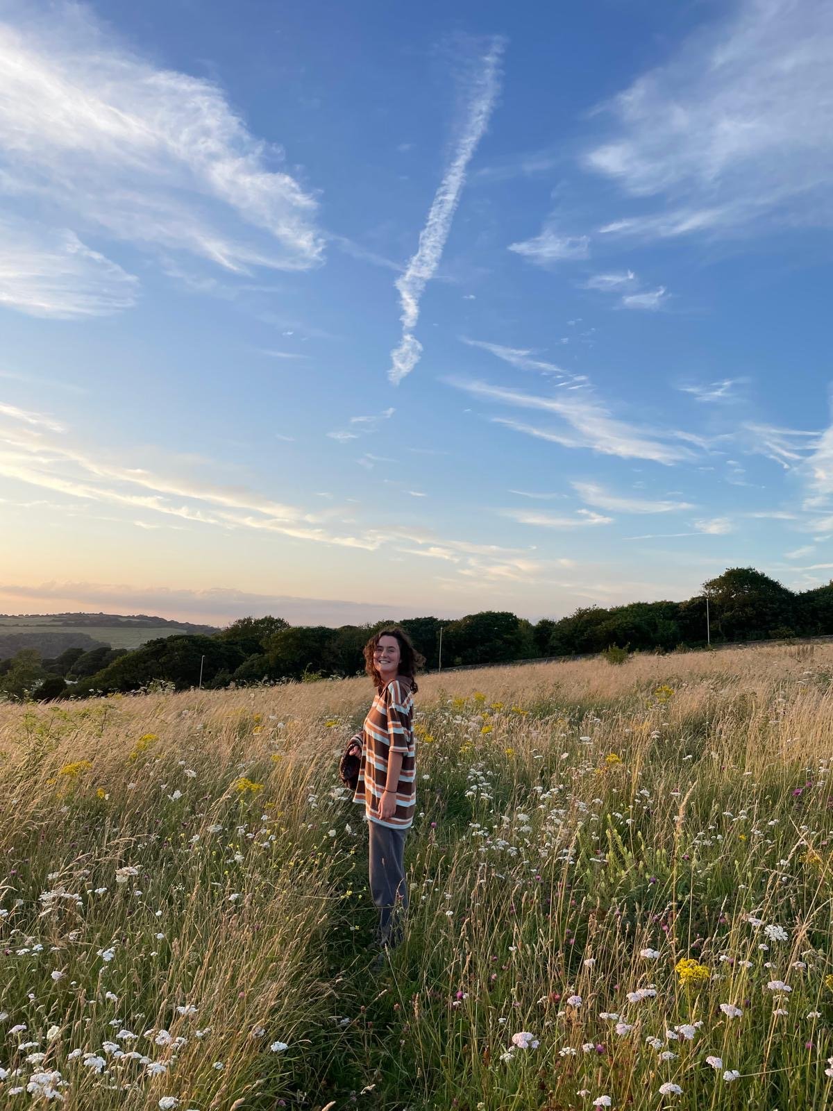 A young woman with curly hair in a striped sweater standing in a grassy field with wildflowers during sunset, under a blue sky with wispy clouds.