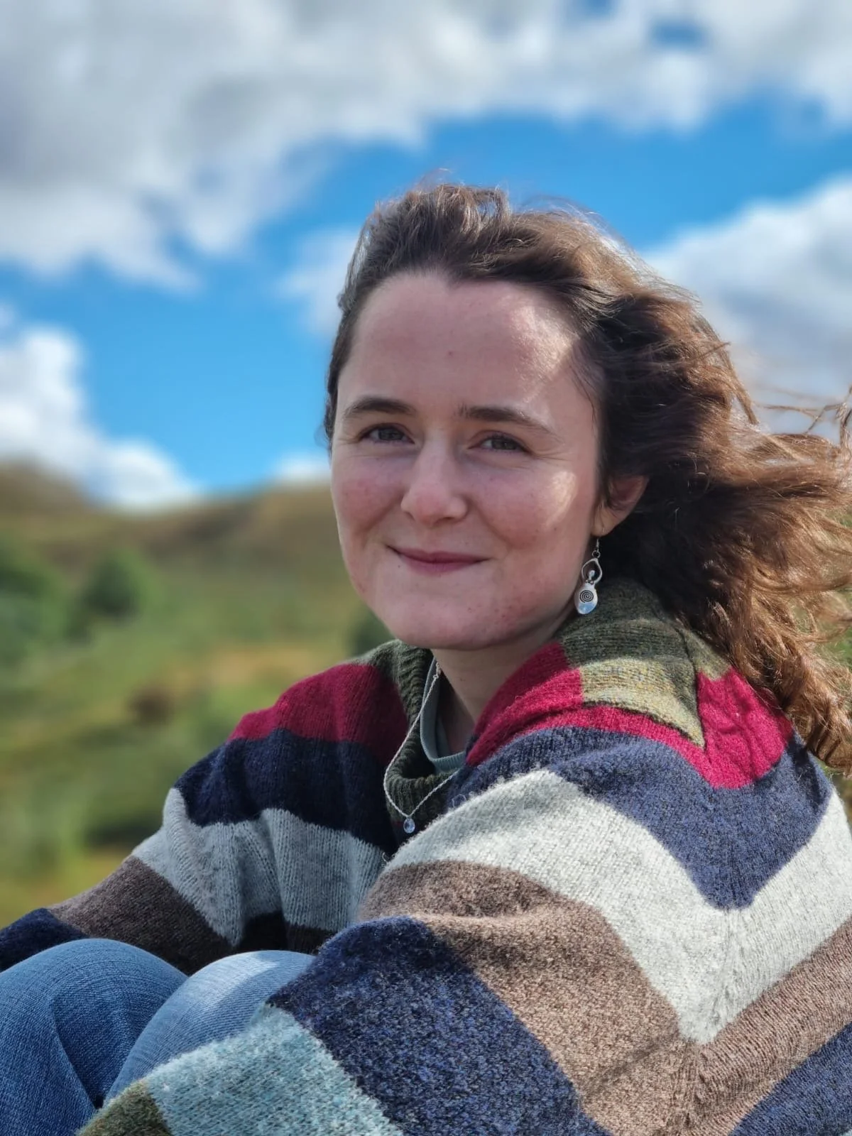 A woman outdoors on a windy day, smiling with her hair blowing, wearing a multicolored striped sweater and earrings, with a grassy hill and blue sky with clouds in the background.