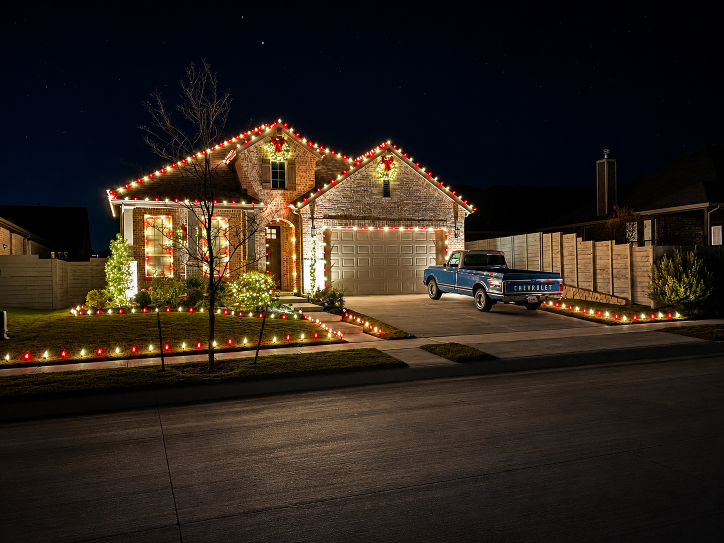 Nighttime view of a house decorated with Christmas lights, including red and white lights outlining the roof, windows, and yard. The house has a brick exterior with a garage and a blue vintage Chevrolet truck parked in the driveway. The yard features
