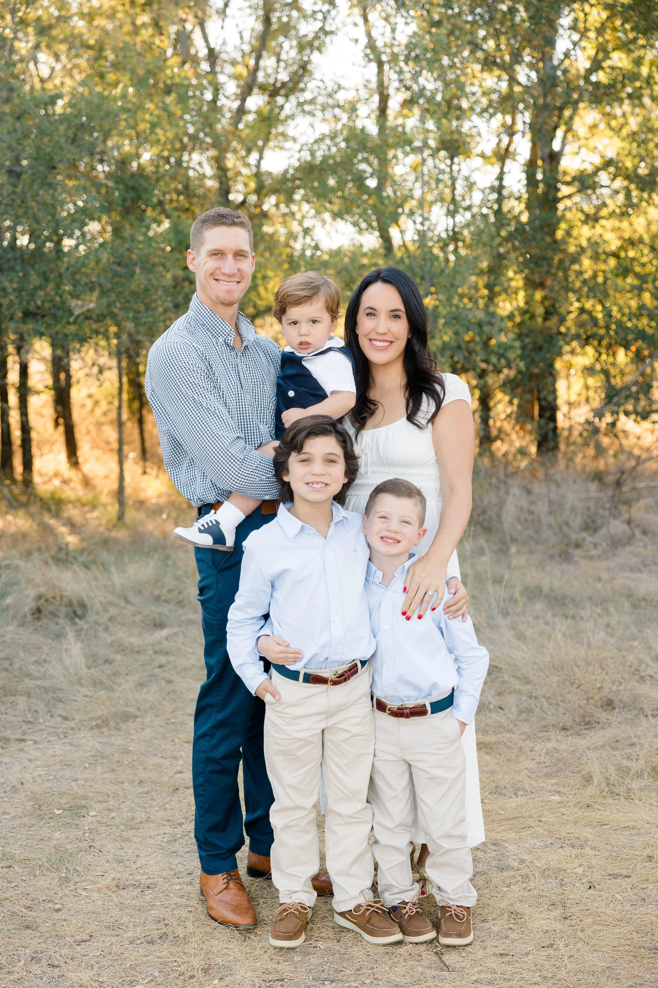 A happy family of six posing outdoors in a wooded area with fall foliage, smiling at the camera. The family consists of a father, mother, and four children, all wearing casual and semi-formal clothing.