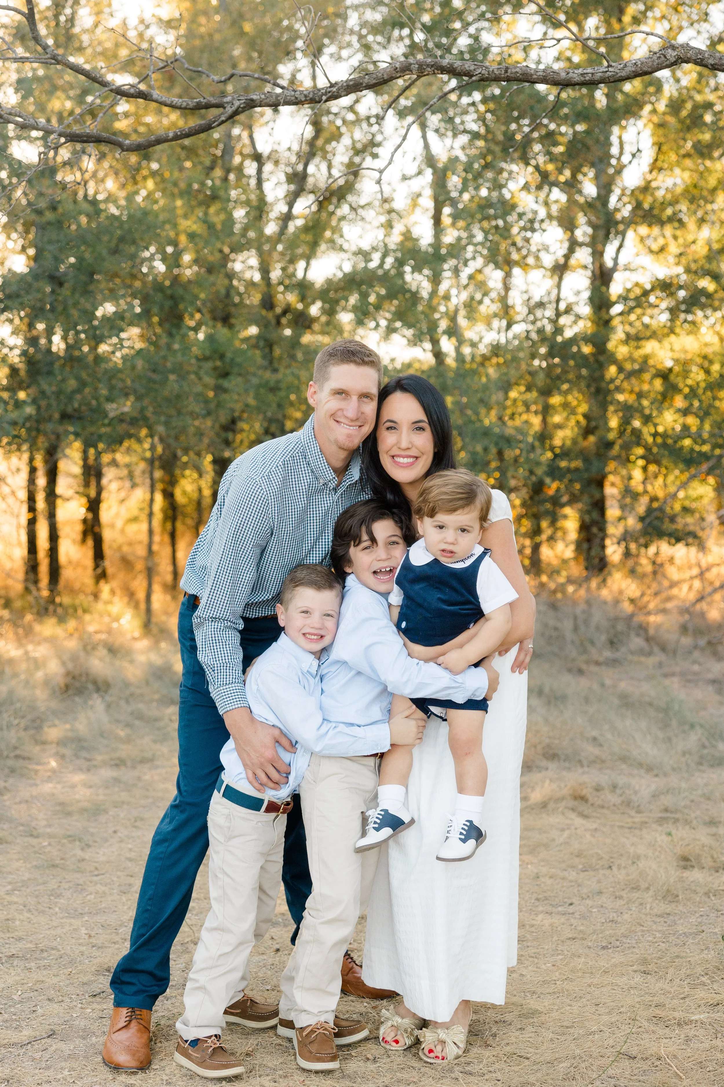 A smiling family of six standing outdoors in a wooded area during golden hour, embracing each other. The family includes two adult parents, three children, and one toddler. The background features trees with autumn foliage and a branch overhead.