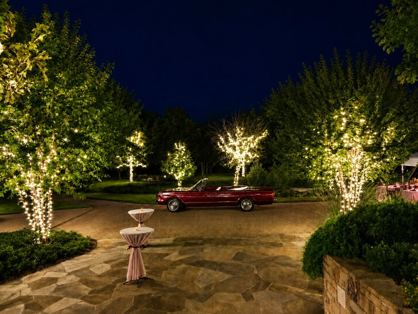Night scene of a lush garden with trees decorated with string lights. A vintage red convertible car is parked on a circular driveway, and two cocktail tables with white cloths and candles are in the foreground.