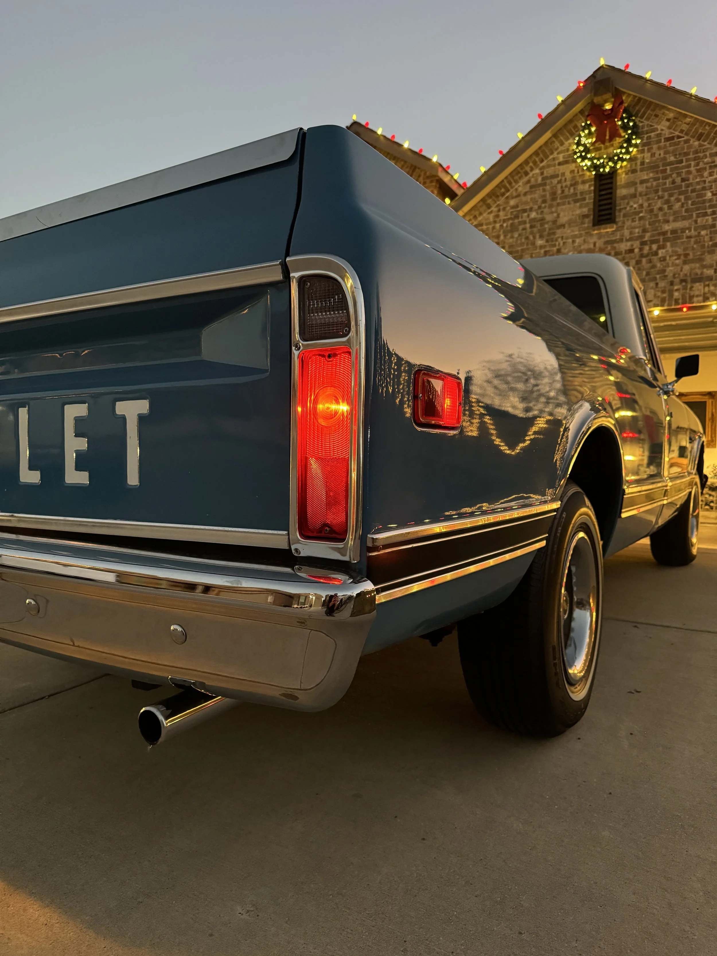 A vintage blue pickup truck parked outside a house decorated with Christmas lights and a wreath in the evening.