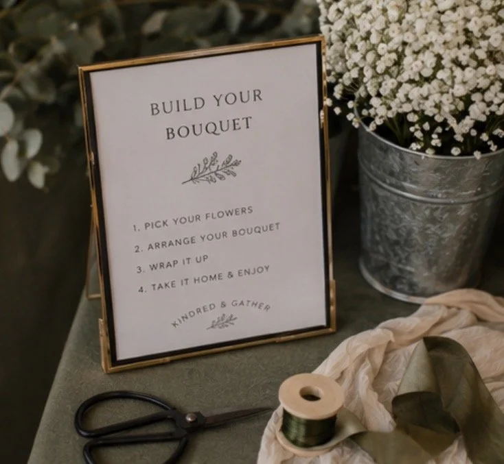Sign with instructions for building a floral bouquet, next to a pair of scissors, spool of green ribbon, and a bucket of white flowers.