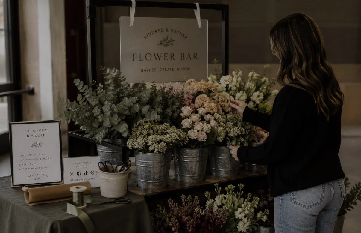 A woman arranging flowers at a flower stand with buckets of various flowers, a sign reading 'Flower Bar,' and instructions on building a bouquet.