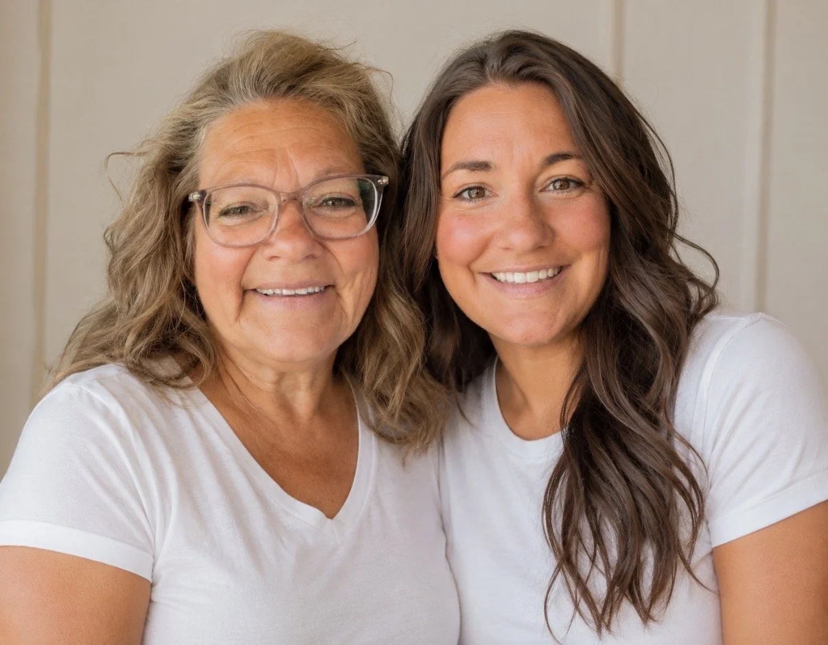 Two smiling women, one older with glasses and curly hair, and one younger with long wavy hair, wearing white t-shirts, standing close together indoors.