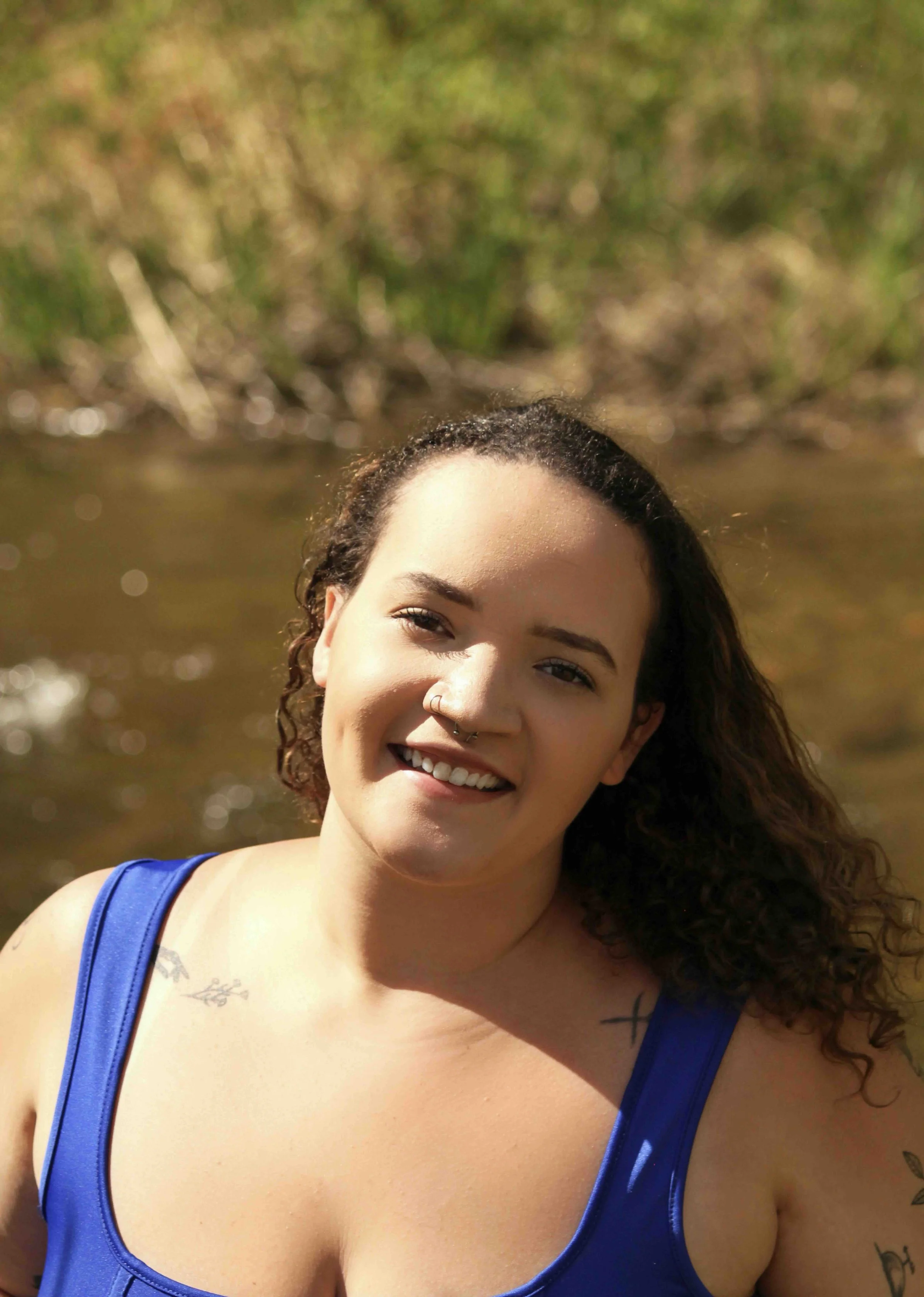 Close-up of a smiling young woman with curly hair, wearing a blue tank top, outdoors near water, with a blurred background of green foliage.