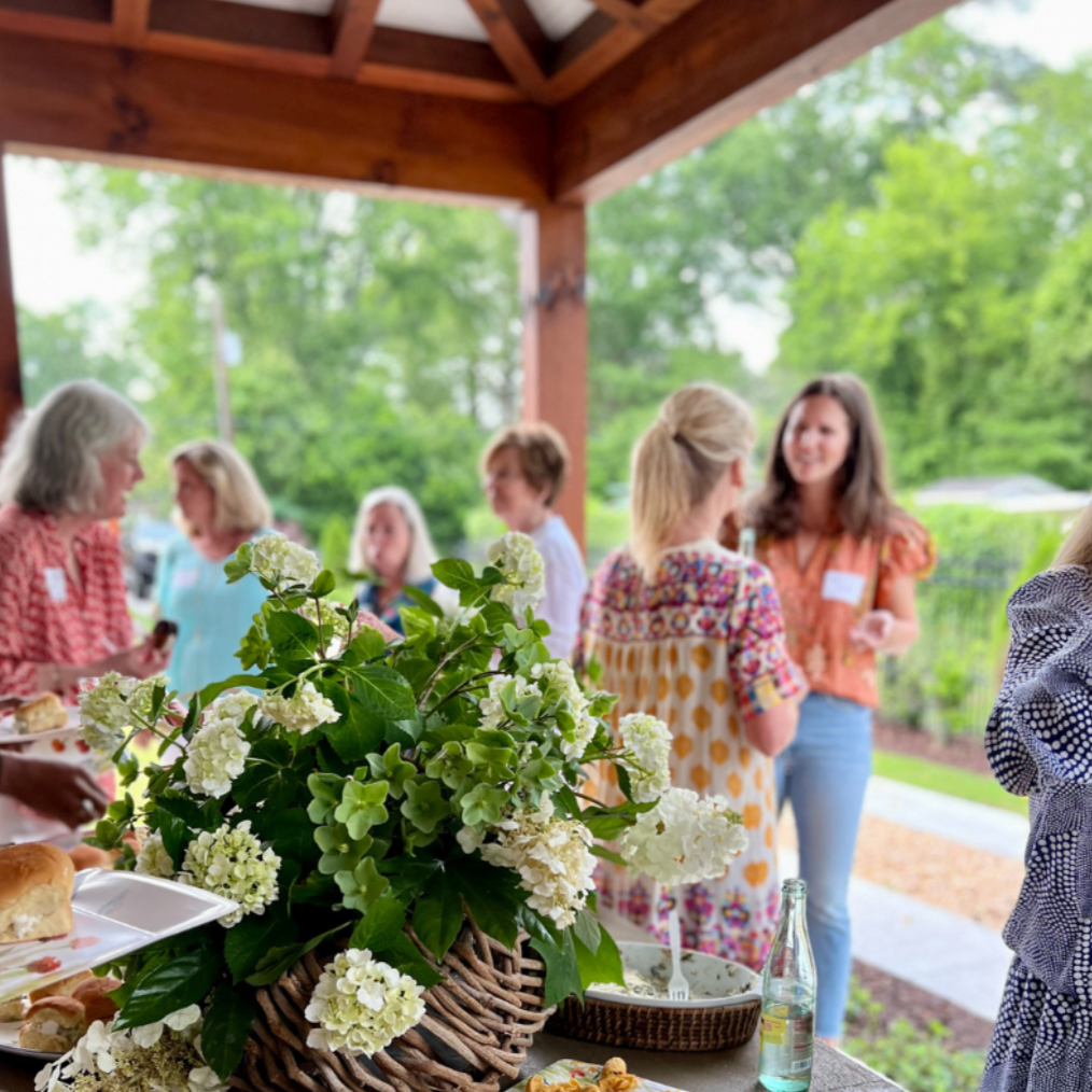 People socializing at an outdoor gathering under a wooden pavilion, with a table of food and a large centerpiece of white hydrangeas in the foreground.