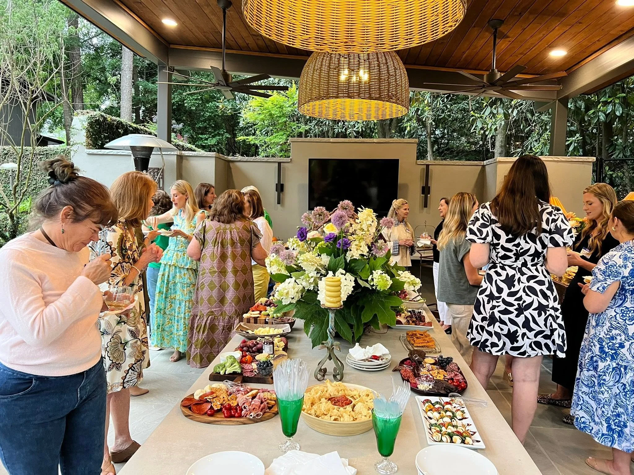 People gathered around a table for our annual Ladies' Night with food at an outdoor gathering. The table has a floral centerpiece and an assortment of dishes, including fruits, appetizers, and desserts.