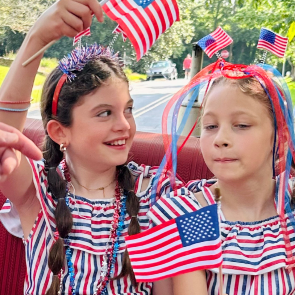 Celebrating America's Independence Day on Riverdale at our annual 4th of July Parade; girls holding small American flags and wearing star-spangled headbands, with flags attached to their costumes.