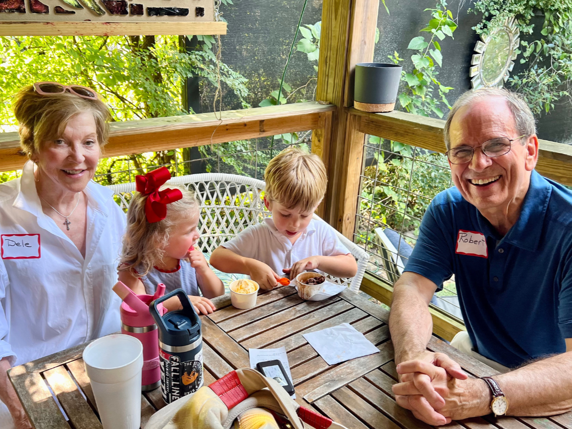 Two kids and two adults, sitting at a wooden table in a sunlit outdoor screened porch with greenery in the background. The adults are smiling, and the children are eating ice cream.