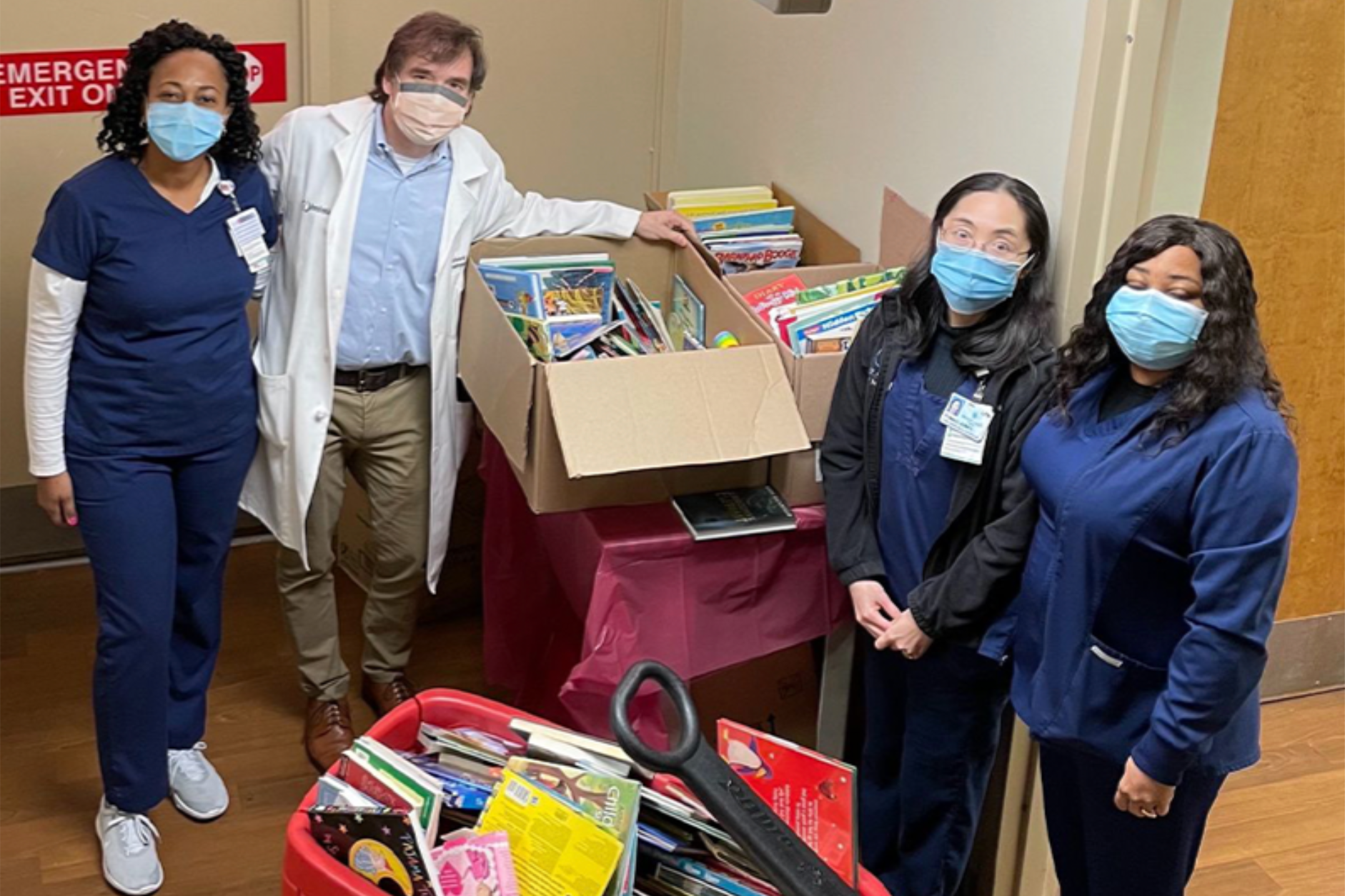 Four healthcare workers, three women and one man, wearing face masks, stand beside tables filled with books and supplies, involved in a book drive or donation event, inside a room with a red emergency exit sign visible in the background.