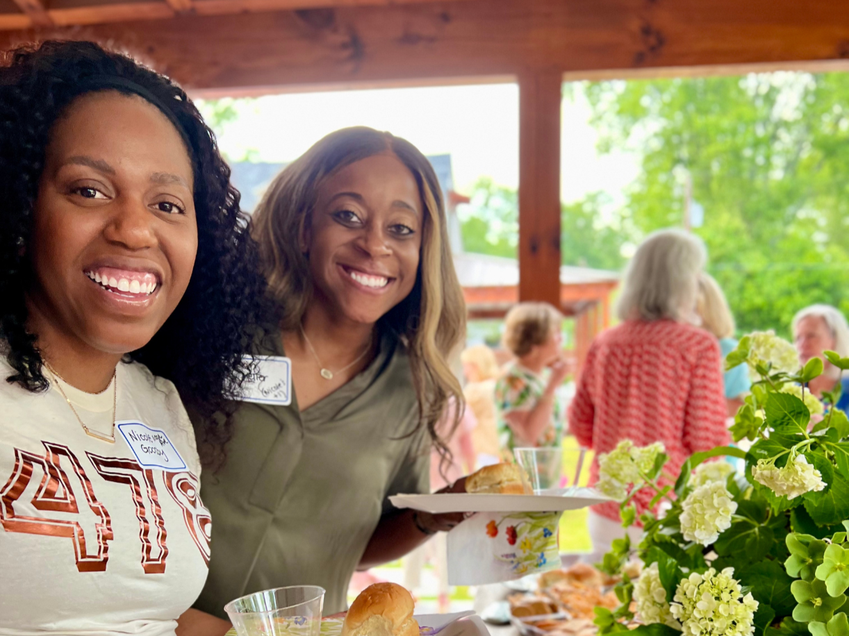 Two smiling women enjoying our annual Ladies' Night - standing at an outdoor gathering, holding plates with food, with other people and greenery in the background.