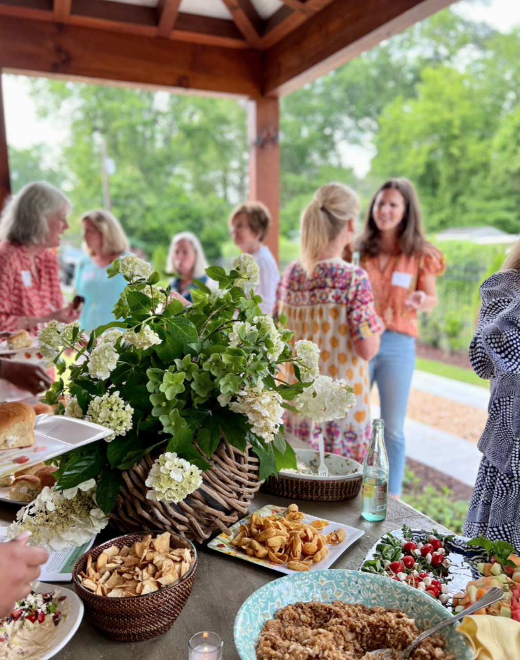 A gathering of women socializing outdoors at a party or gathering under a wooden pavilion, with a table of food including a large floral centerpiece, chips, salad, and other dishes, with green trees in the background.