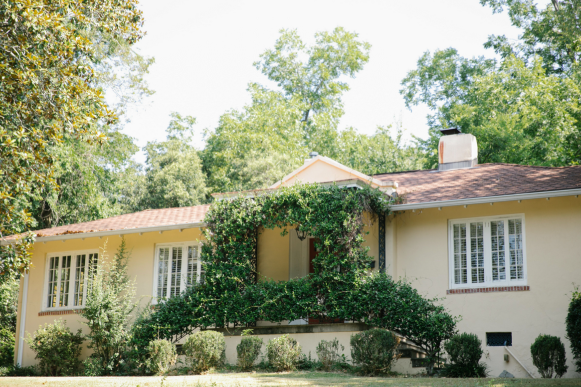 Front view of a yellow house with a red tile roof, surrounded by green trees and shrubs, with a ledge and a balcony covered in climbing greenery.