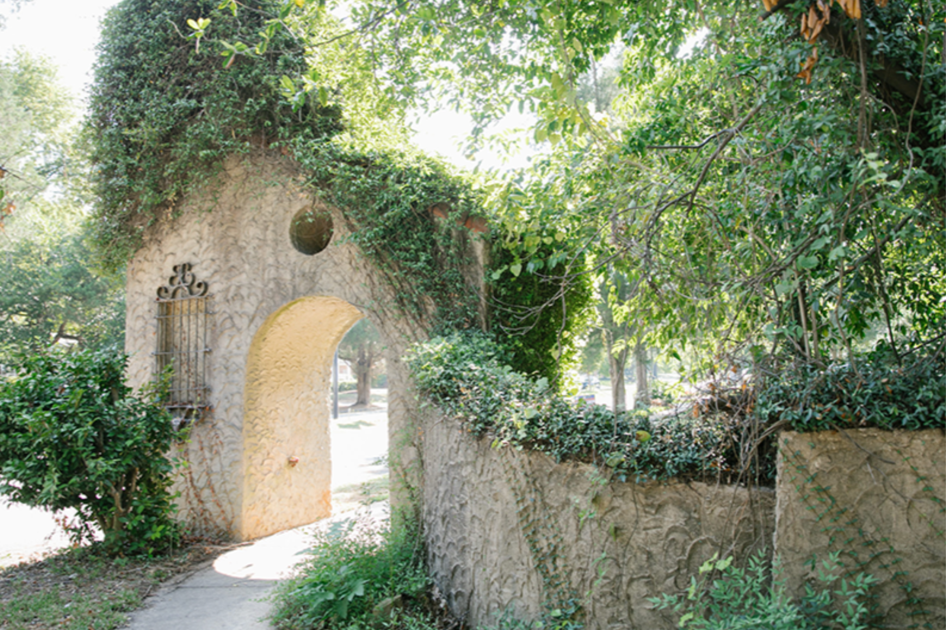 A picturesque entryway onto The Prado through an archway covered with vines and greenery, with a textured stone wall and a wrought iron gate on the left side.