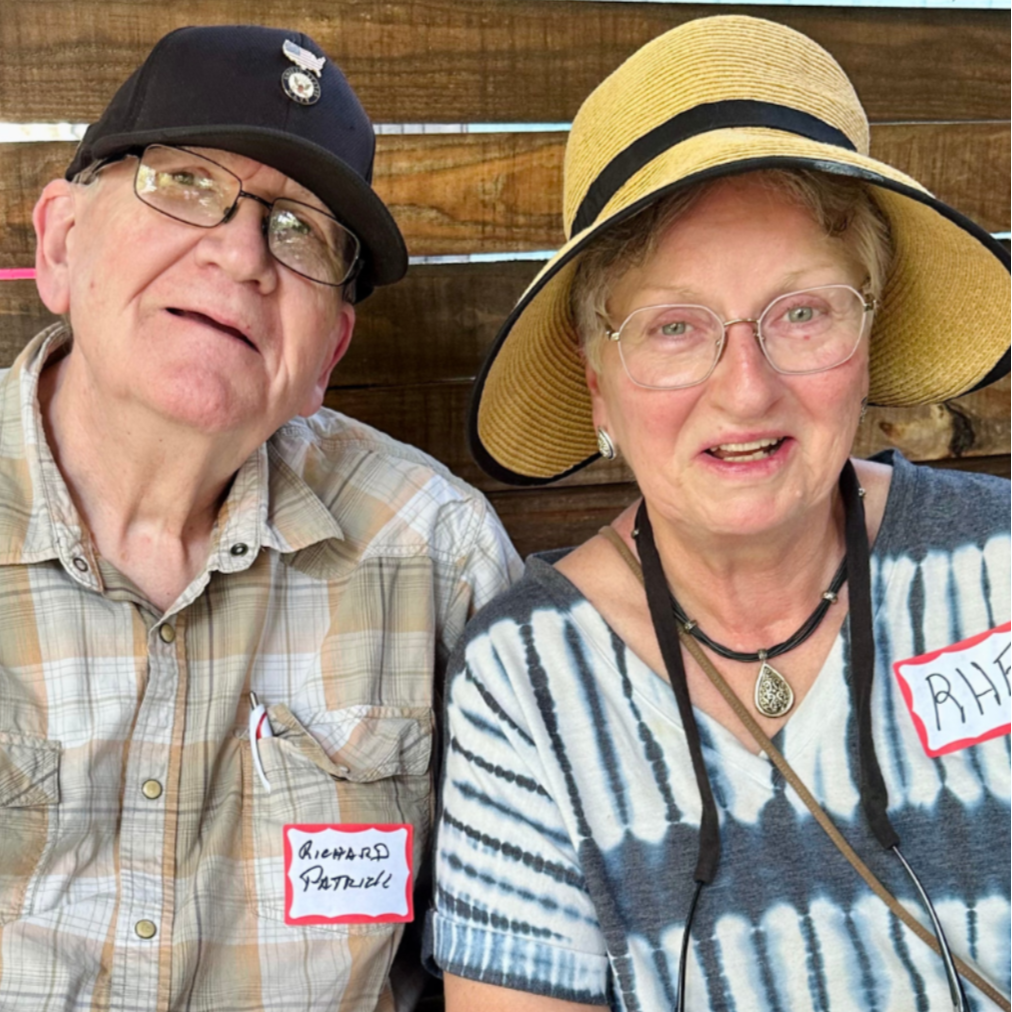 A man and woman sitting next to each other outside, both wearing glasses and smiling. The man has a black cap with pins and a plaid shirt, while the woman has a wide-brimmed straw hat, a striped shirt, and a necklace.