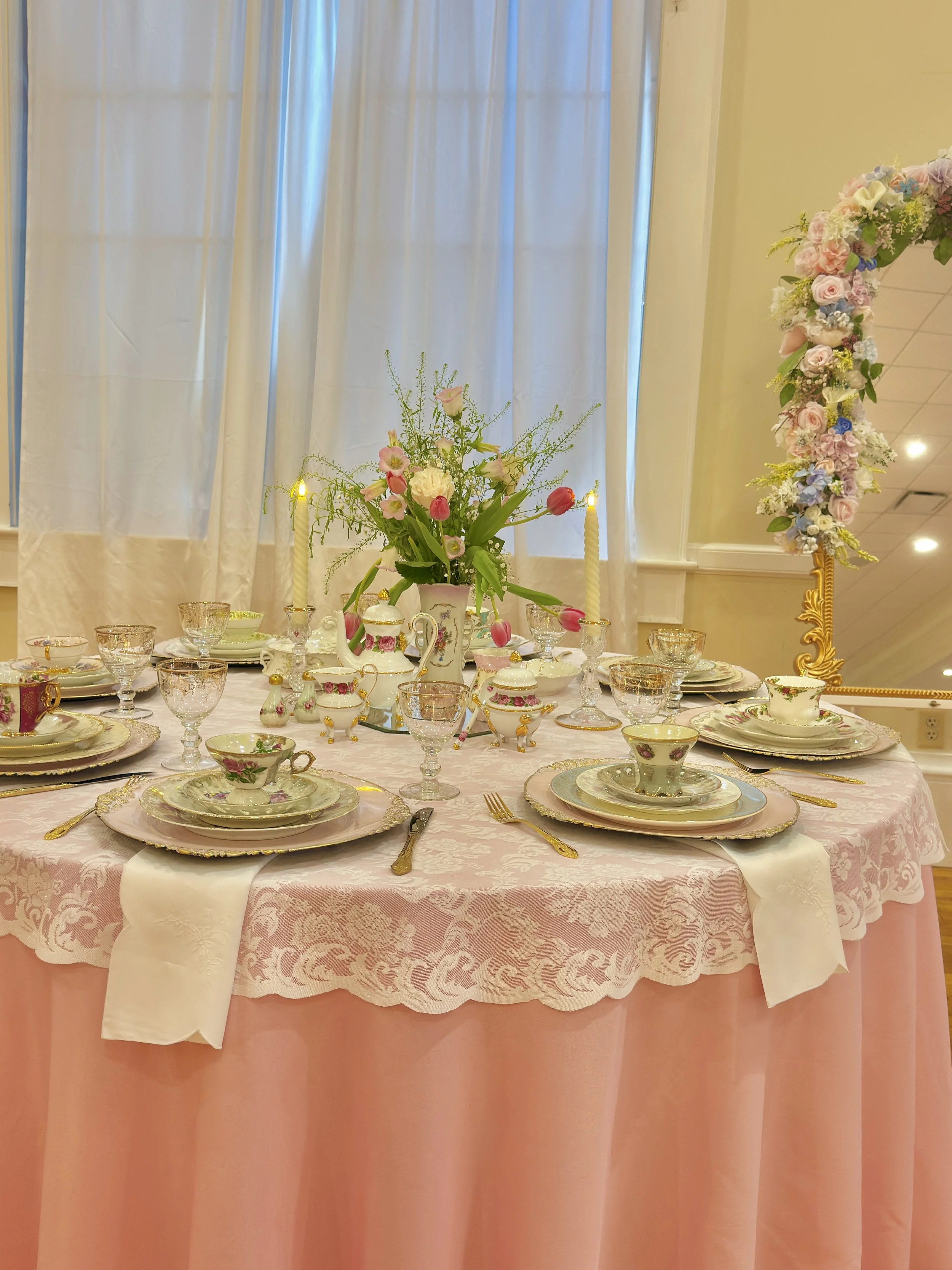 Elegant round table set with fine china, glassware, gold utensils, and a floral centerpiece, with pink tablecloth and a pink curtain backdrop.