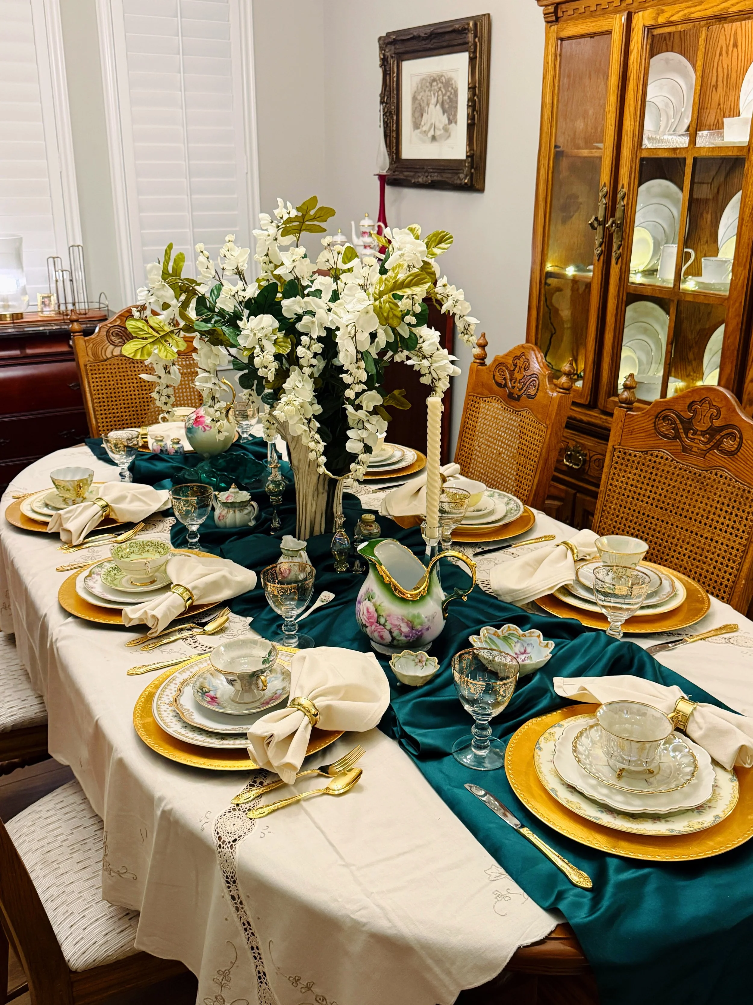 A dining table set for a formal meal with fine china, glassware, gold utensils, and cloth napkins. The centerpiece features a large bouquet of white flowers in a gray vase with a dark cloth runner underneath. In the background, there are wooden chair