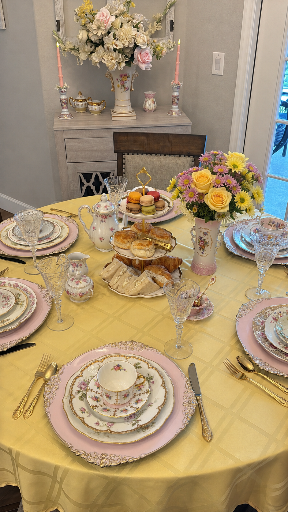 A decorated table set for a special occasion with fine china, crystal glasses, and gold flatware, featuring a centerpiece with yellow roses and pink daisies, and a tiered plate with pastries and macarons in the background.