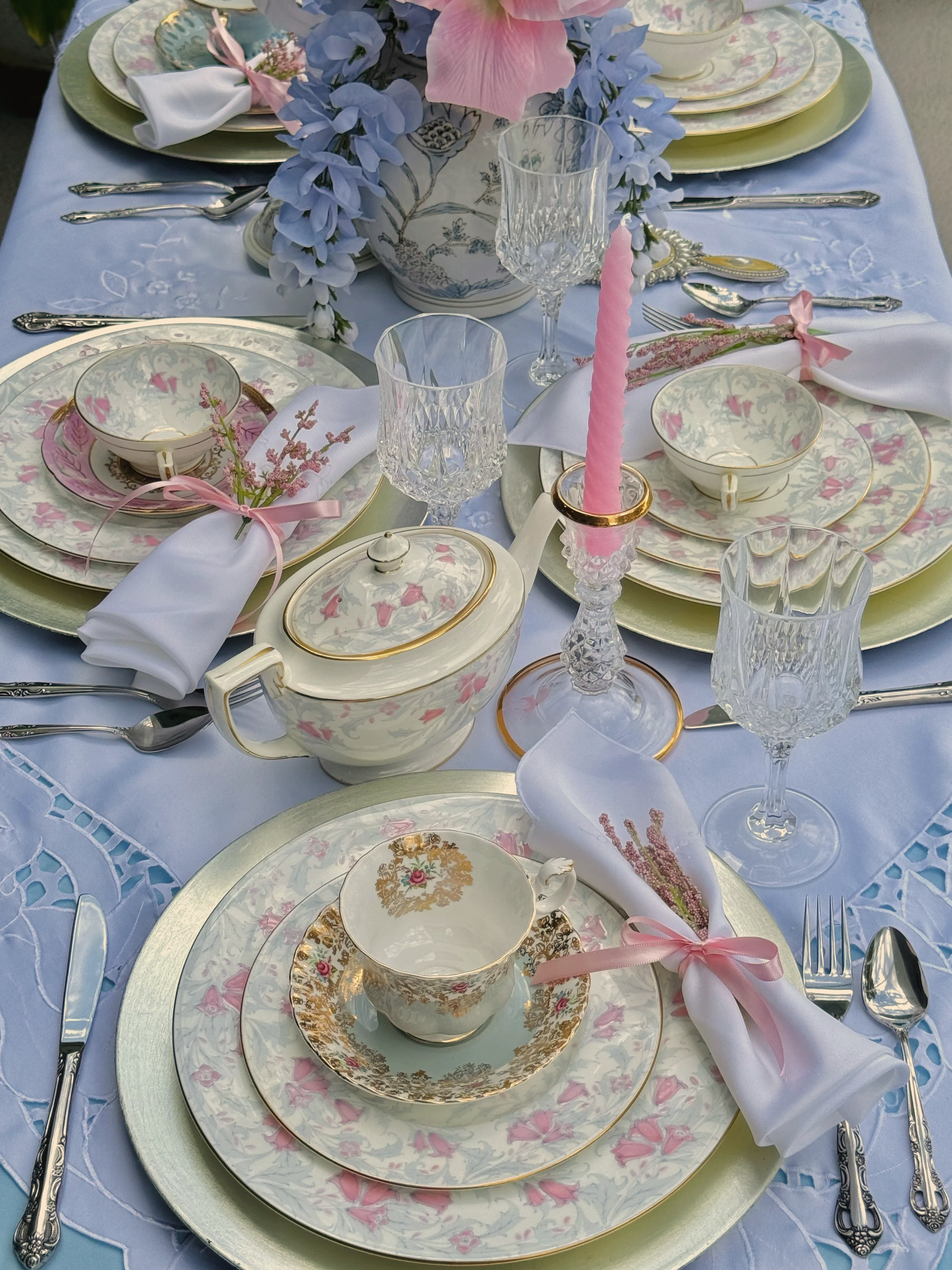 An elegantly set dining table with floral china, crystal glasses, silverware, white napkins tied with pink ribbons, pink candles, and a floral centerpiece with blue and pink flowers, all on a light blue tablecloth.