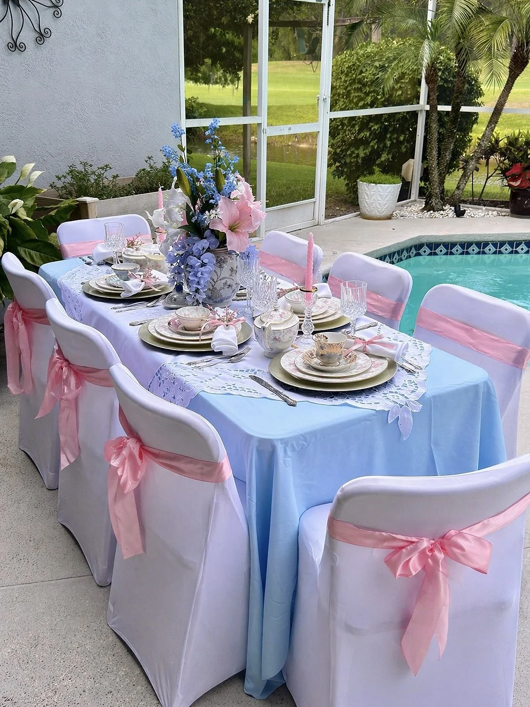 A round outdoor dining table set for a celebration, with white chairs decorated with pink sashes, pastel-colored dinnerware, and a floral centerpiece, next to a swimming pool on a patio with a screened enclosure and lush greenery.
